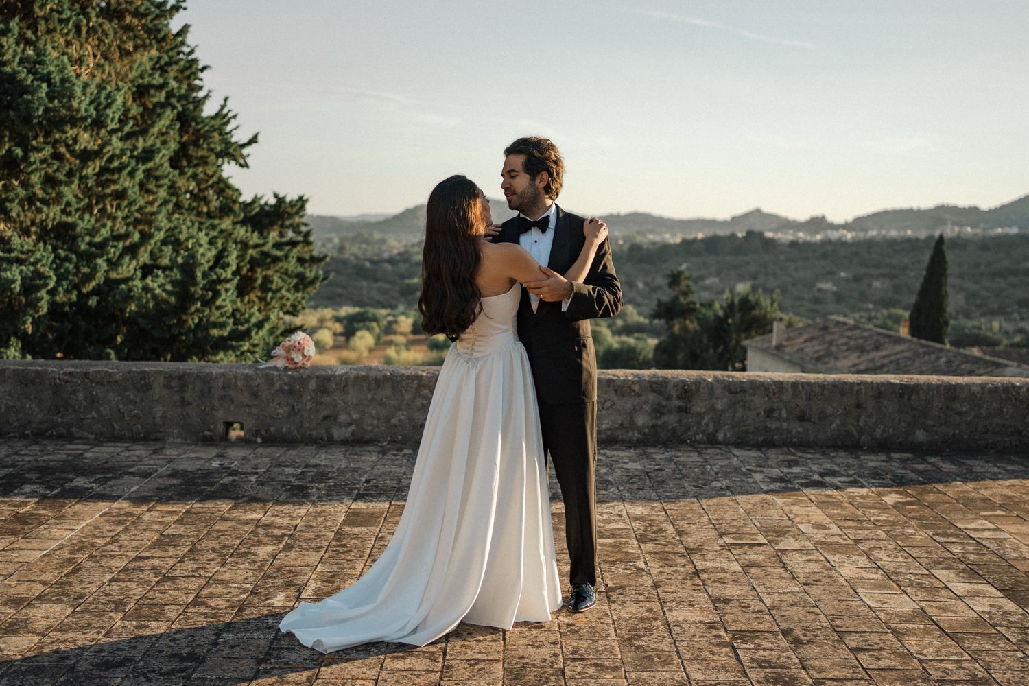A romantic couple embraces during their wedding photoshoot on a rustic stone terrace overlooking scenic mountains.