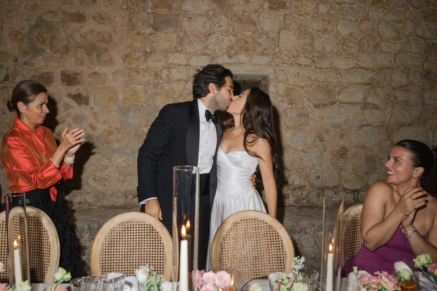 Wedding guests seated at a long rustic dining table with stone walls in the background during an elegant reception.