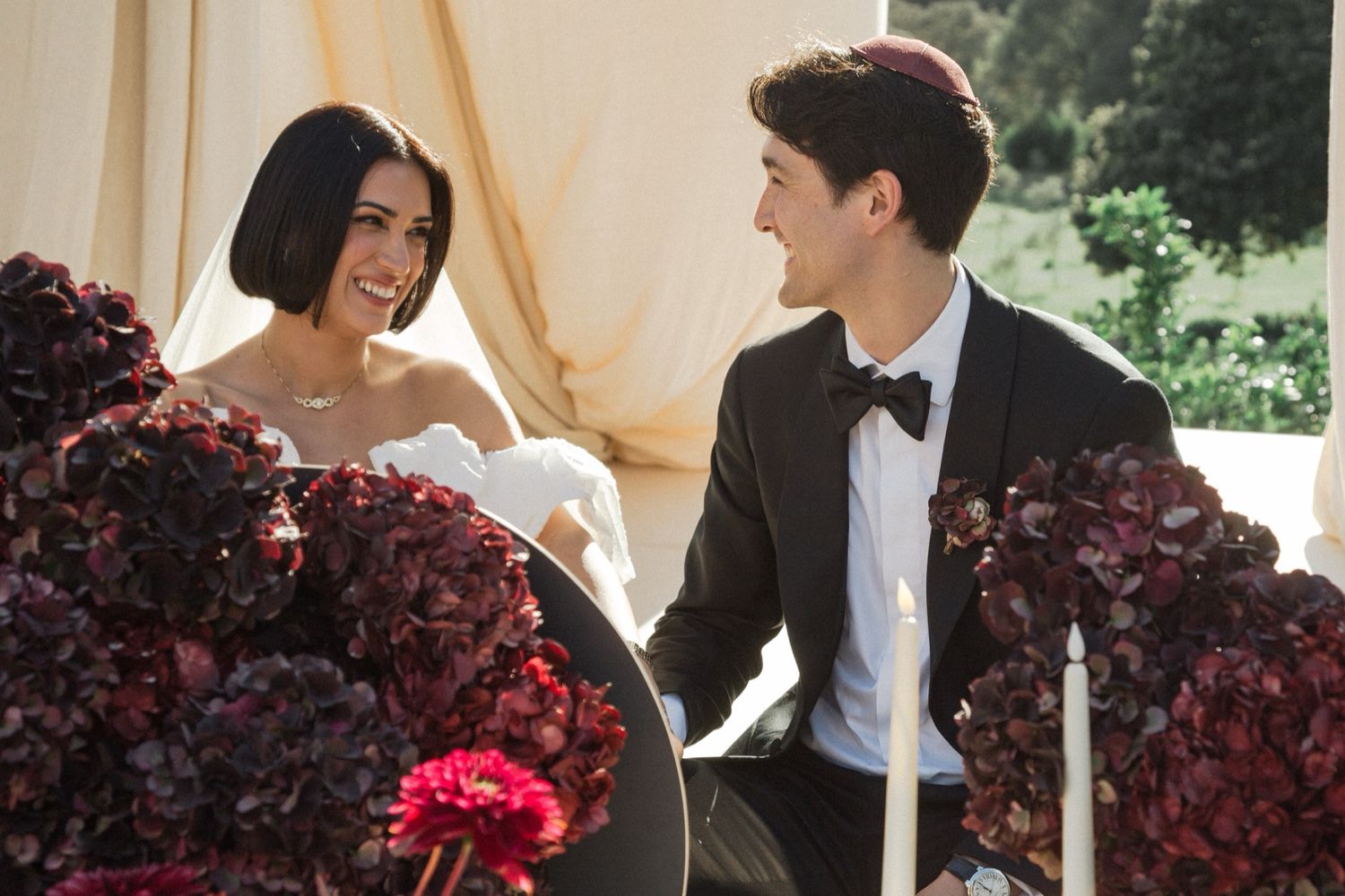 Elegant wedding couple shares a joyful moment with deep burgundy floral arrangements in an outdoor venue.