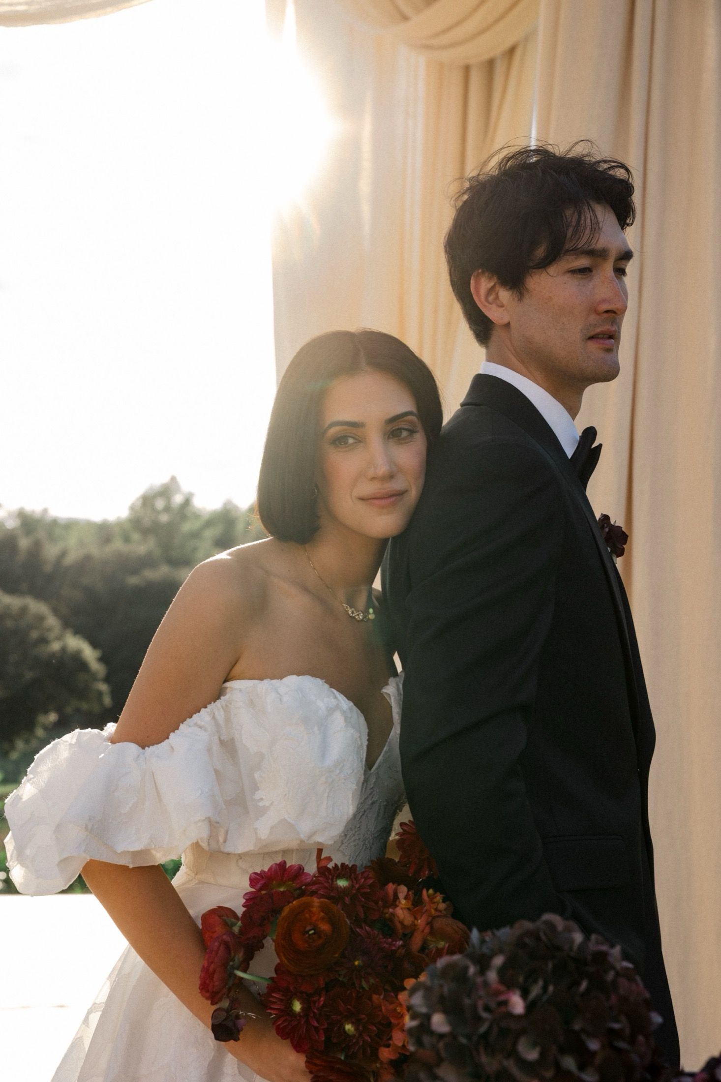 A bride in a white strapless gown stands with her groom in a black tuxedo during an outdoor wedding ceremony.
