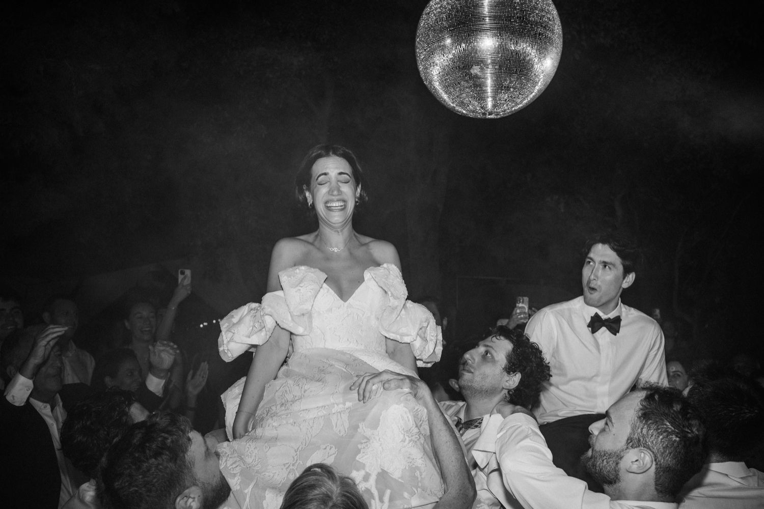 A disco ball glows above a lively dance party scene with people celebrating in formal attire in black and white.