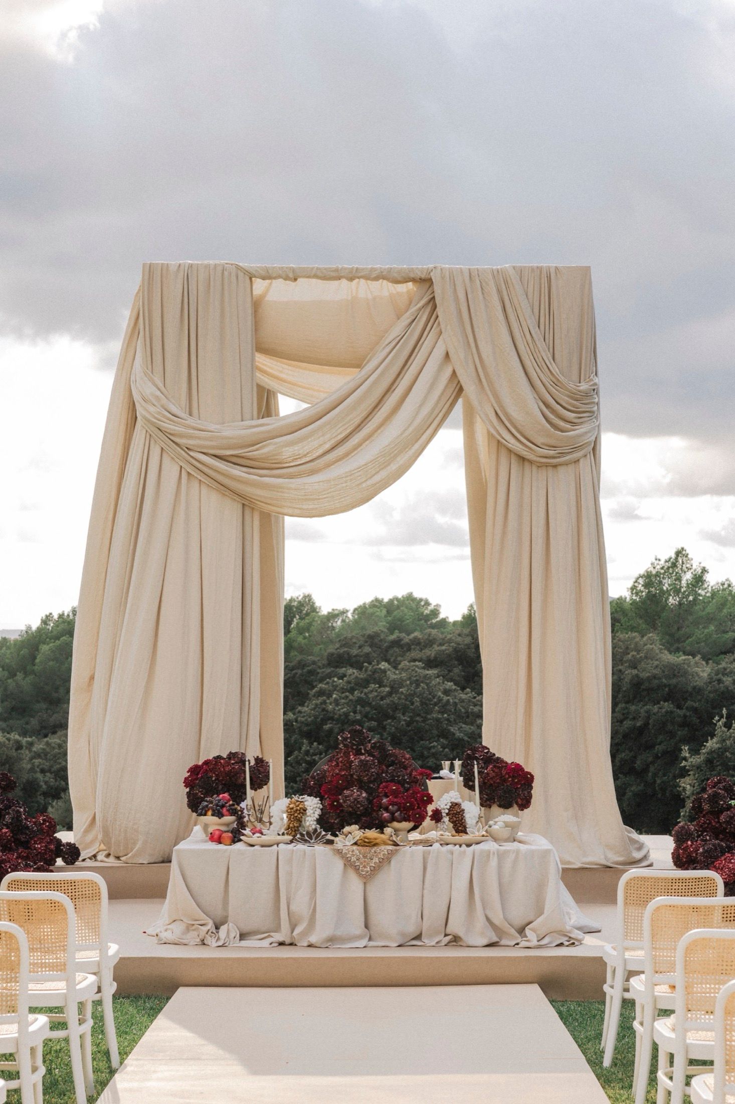 Elegant outdoor wedding reception setup with flowing cream drapes and red floral centerpieces on long white tables.