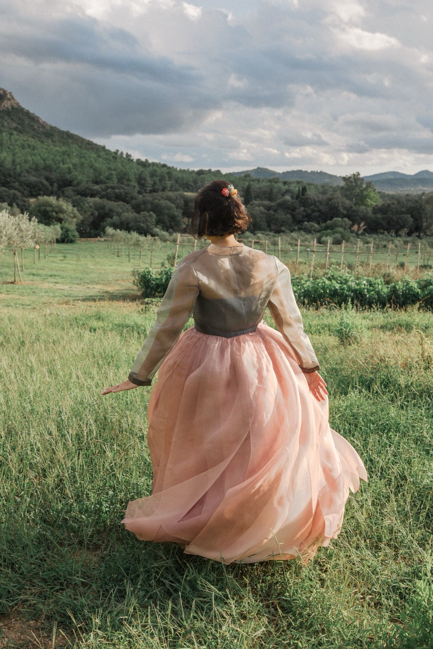 A person in a blush pink gown twirls in a green meadow with mountains in the background.