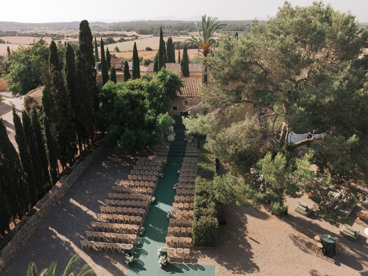 Outdoor wedding ceremony setup with rows of chairs and a green carpet aisle surrounded by palm trees and stone walls.