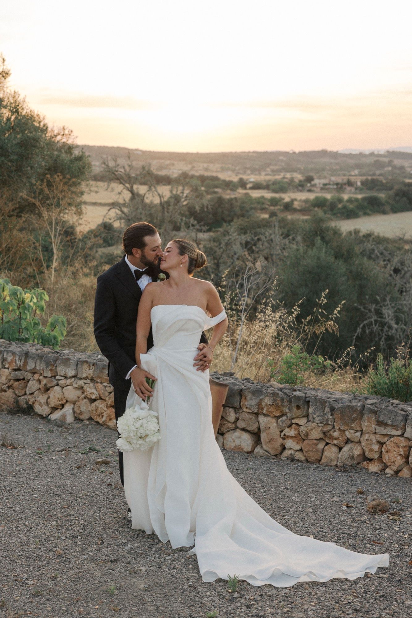 Elegant couple in wedding attire poses against rustic stone wall and rolling hills at golden hour.