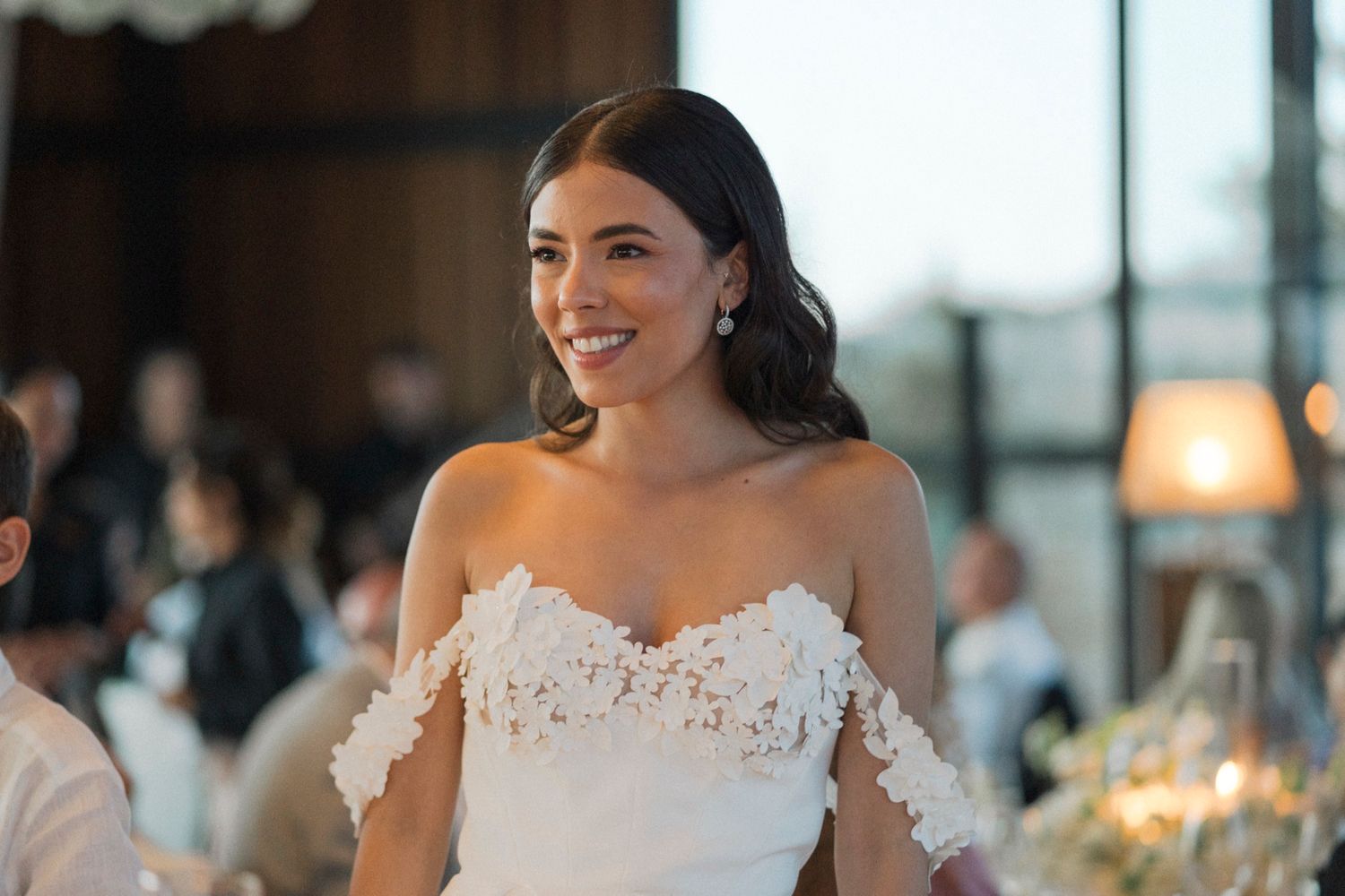 Smiling bride in white off-shoulder lace wedding gown at elegant indoor reception venue.