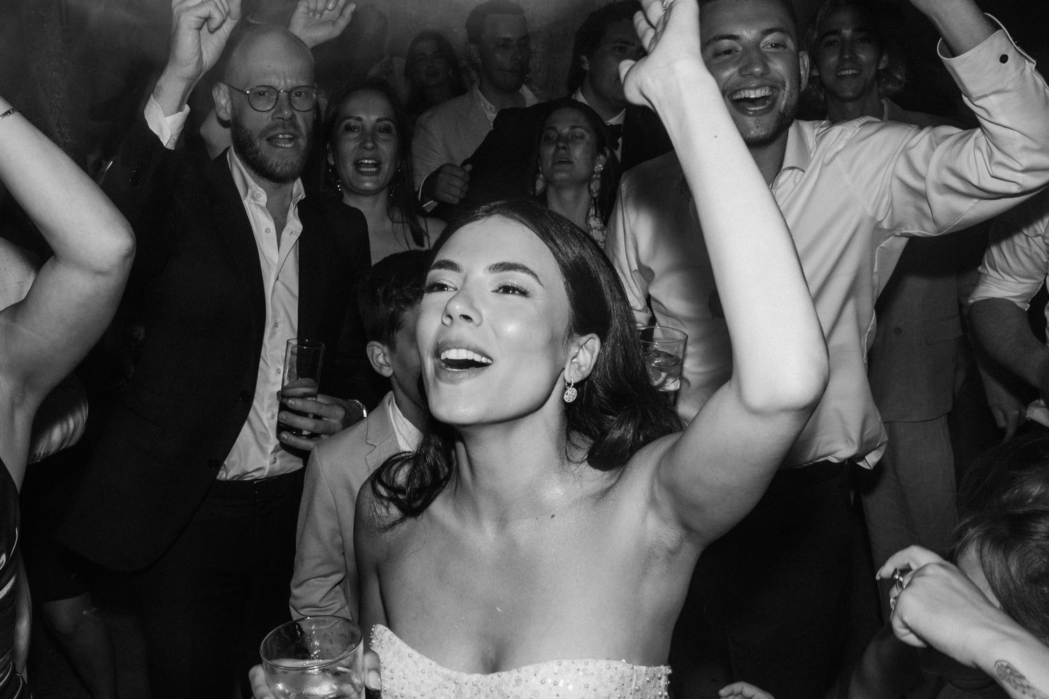Black and white photo of a joyful bride dancing with arms raised at a lively wedding reception.