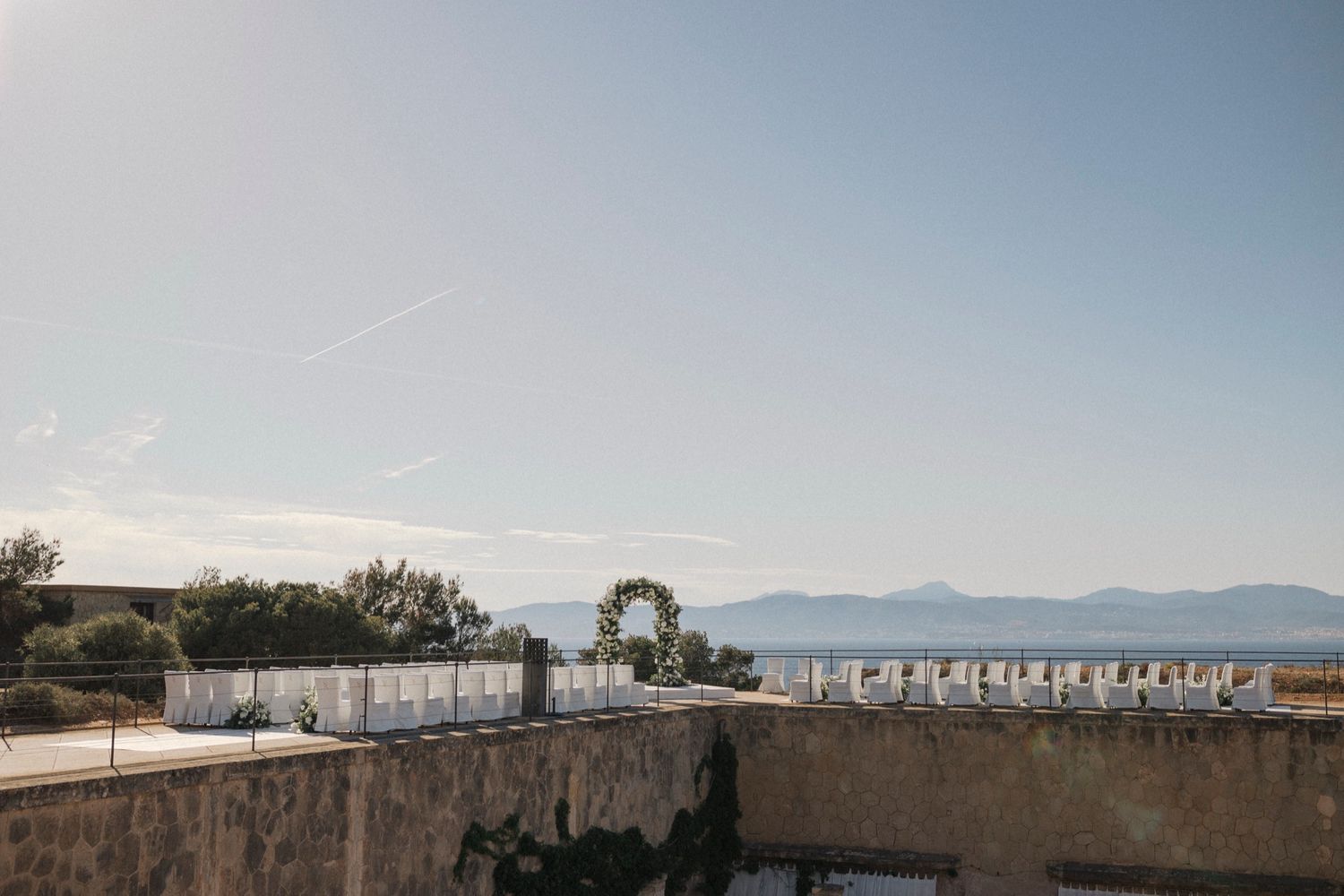 Outdoor wedding ceremony setup with floral arch overlooking a serene sea and mountain backdrop under a clear sky.