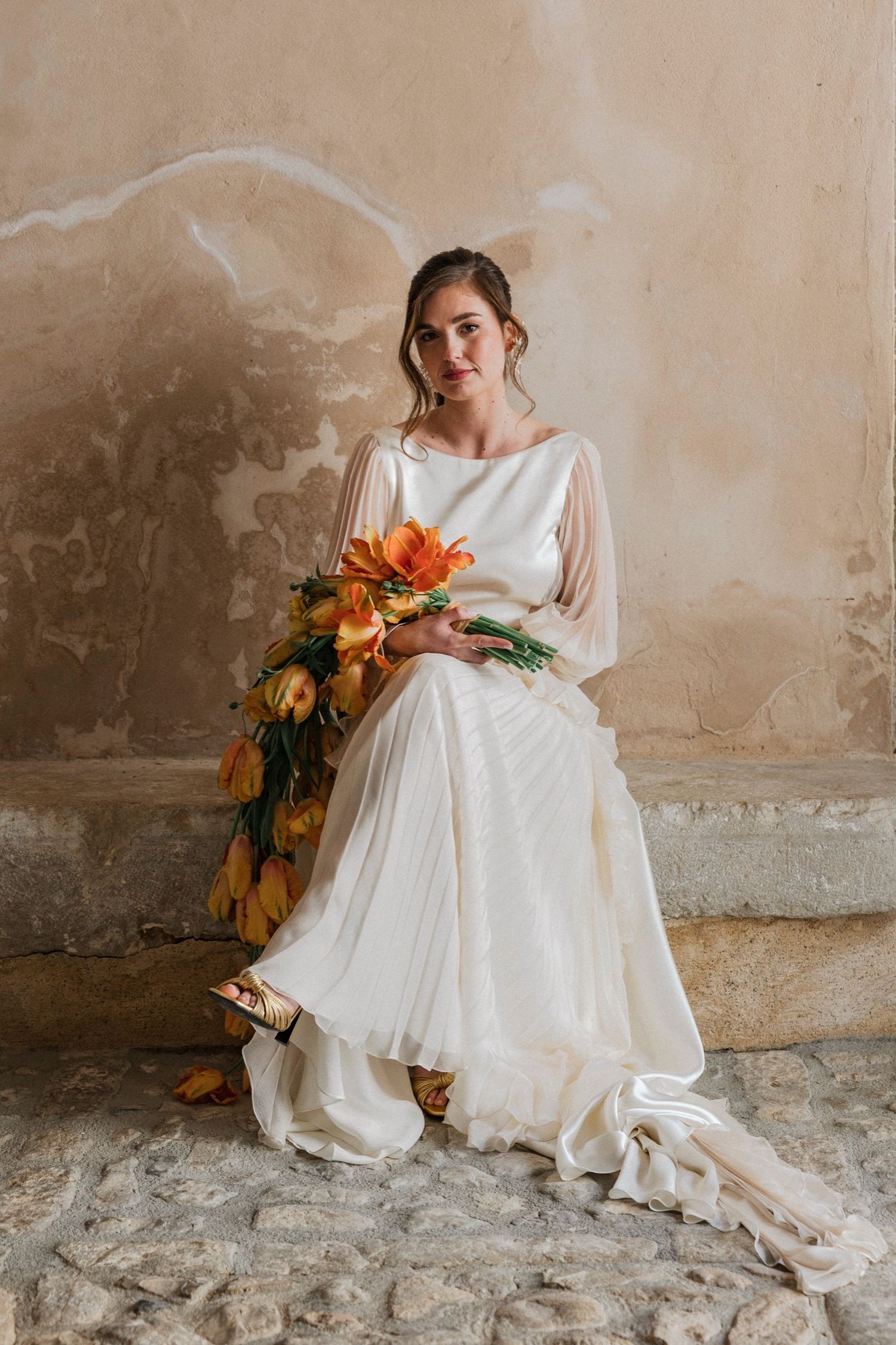 Elegant bridal portrait featuring a flowing white dress and vibrant orange lily bouquet against a textured beige wall.