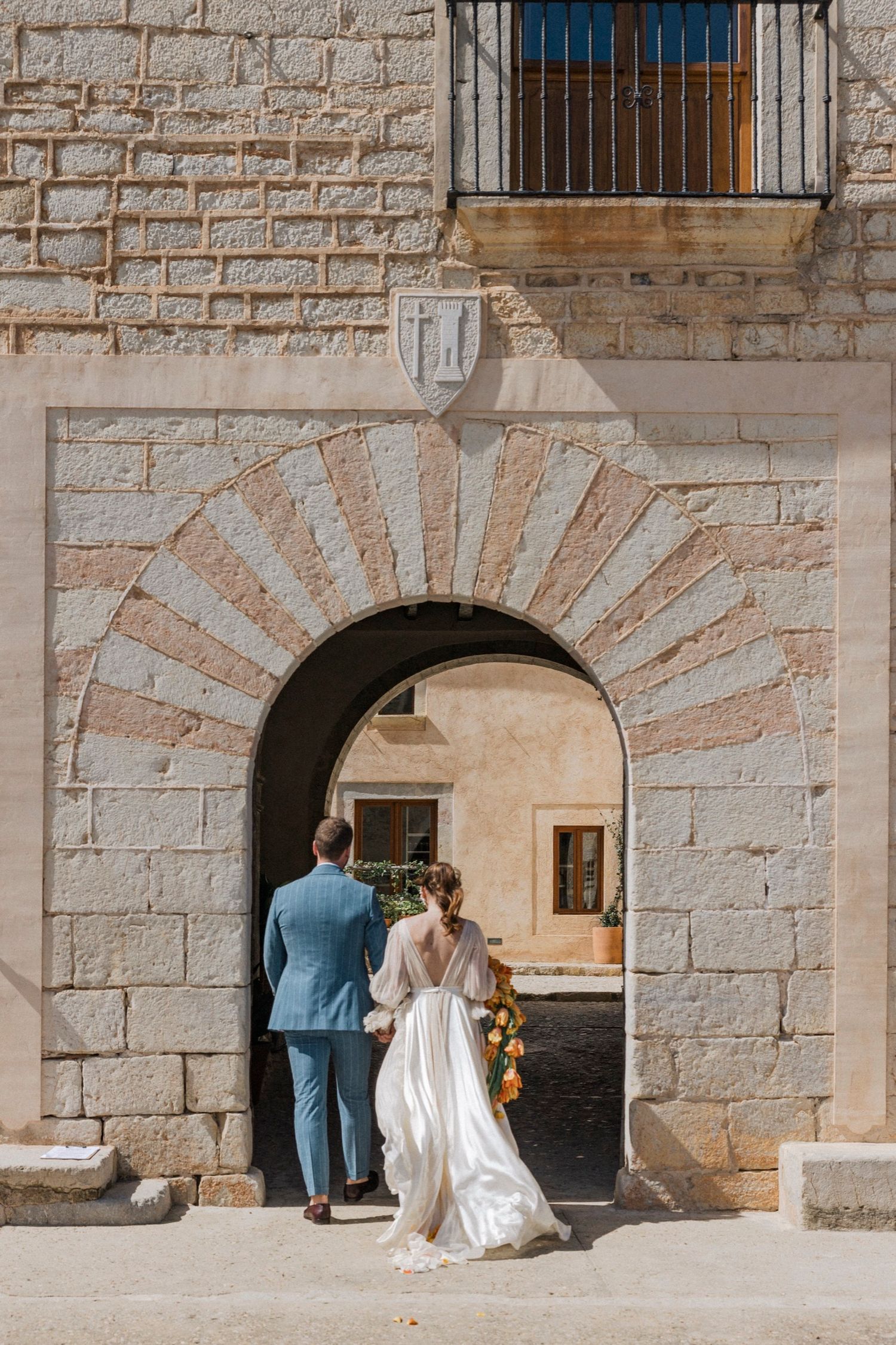 A couple in wedding attire walking through a stone archway with sunburst pattern bricks in a historic building.