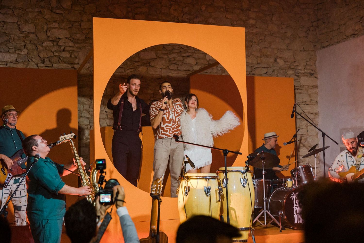 Band members silhouetted against an orange backdrop during a live performance on a dimly lit stage.