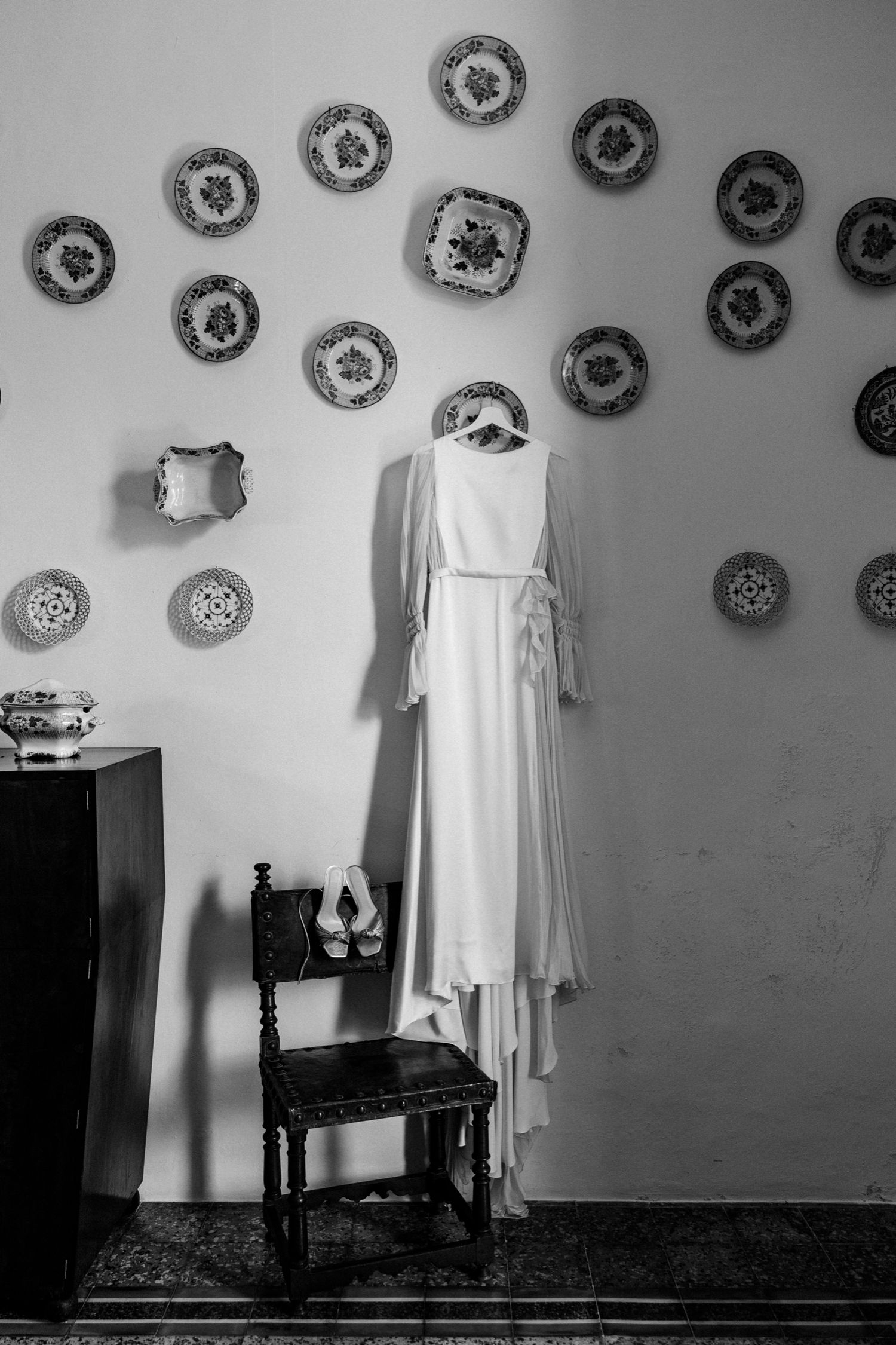 A vintage white dress hangs on a wall decorated with ornate decorative plates in black and white photography.