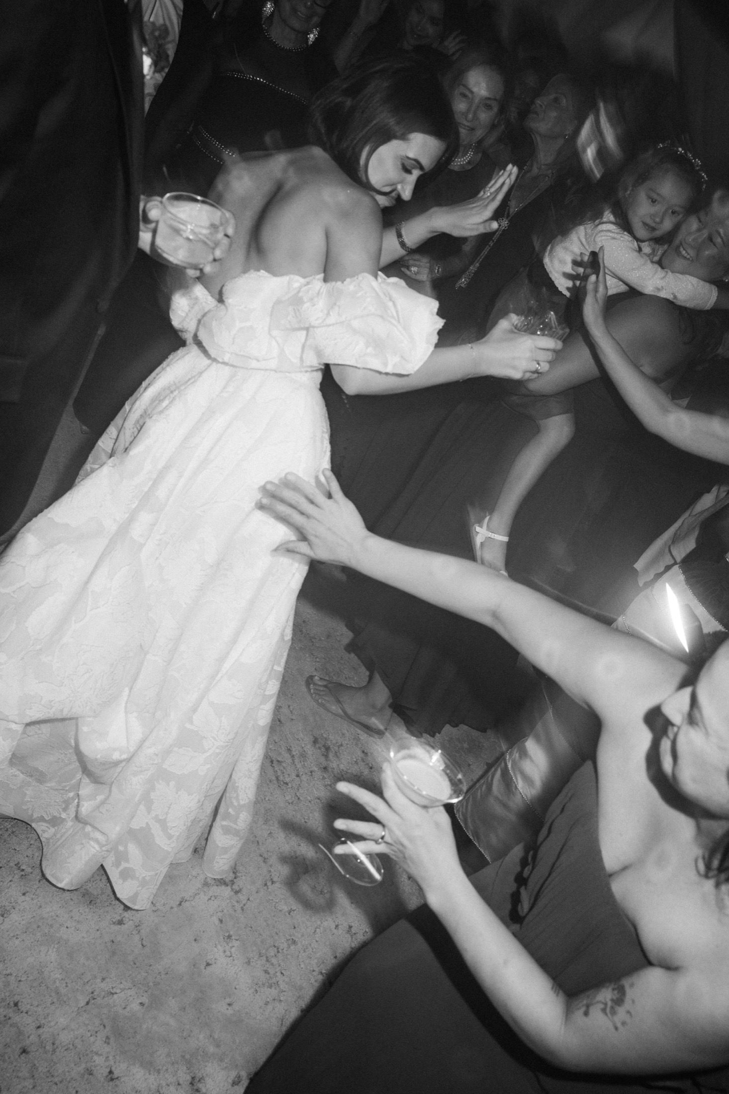 A bride in a white dress dances energetically at a wedding reception while guests surround her on the dance floor.