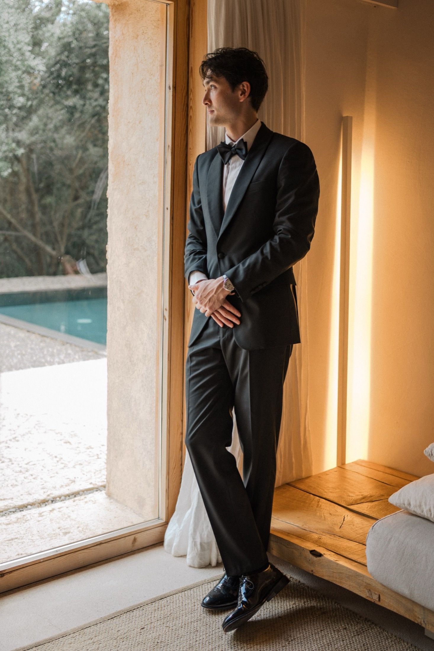 A person in a classic black tuxedo and bowtie stands by a window overlooking a pool in warm lighting.