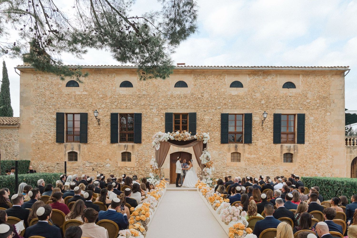 Outdoor wedding ceremony setup with rows of white chairs facing a rustic stone building on a sunny day.