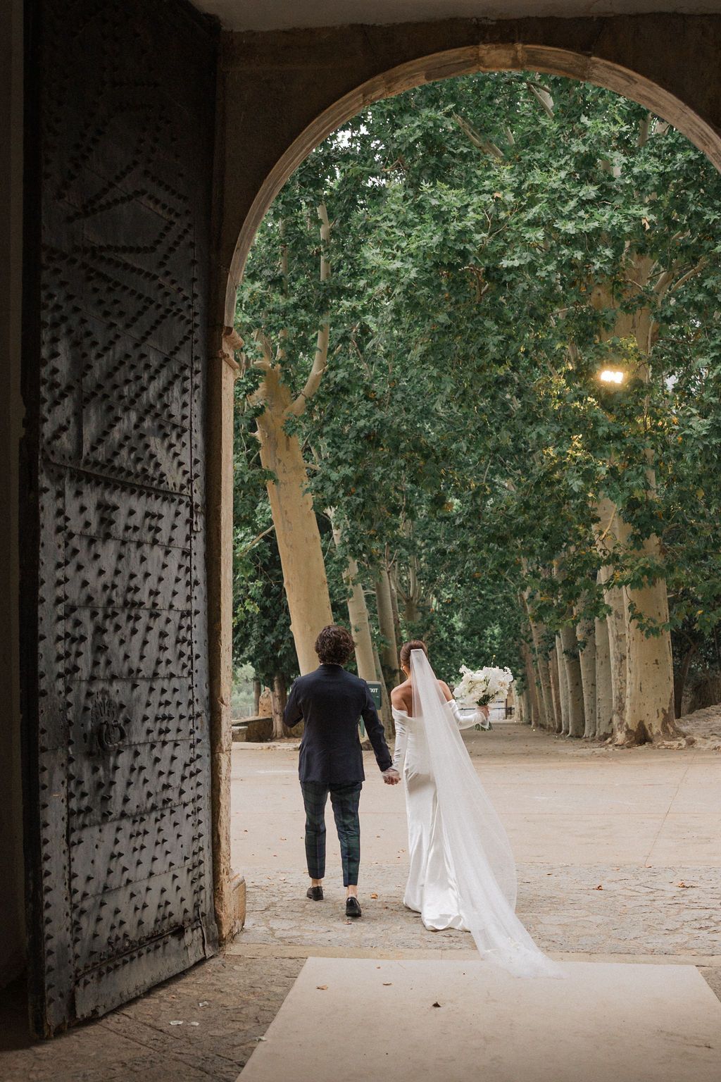 Newlyweds walk through arched passageway lined with trees and ornate metal doors.