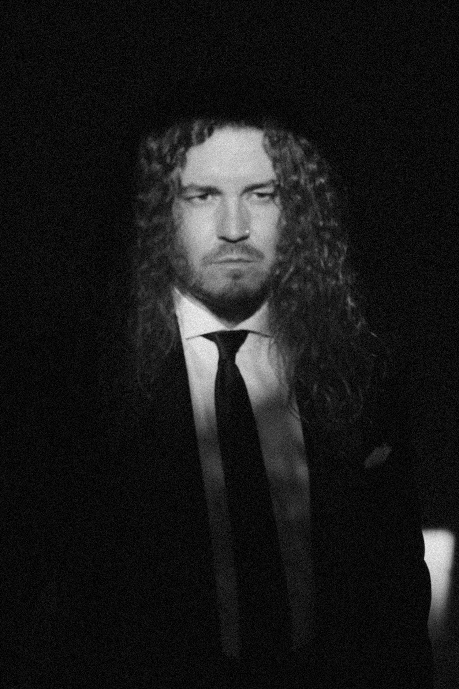 A dramatic black and white portrait of a person with long curly hair wearing a suit and tie against a dark background.