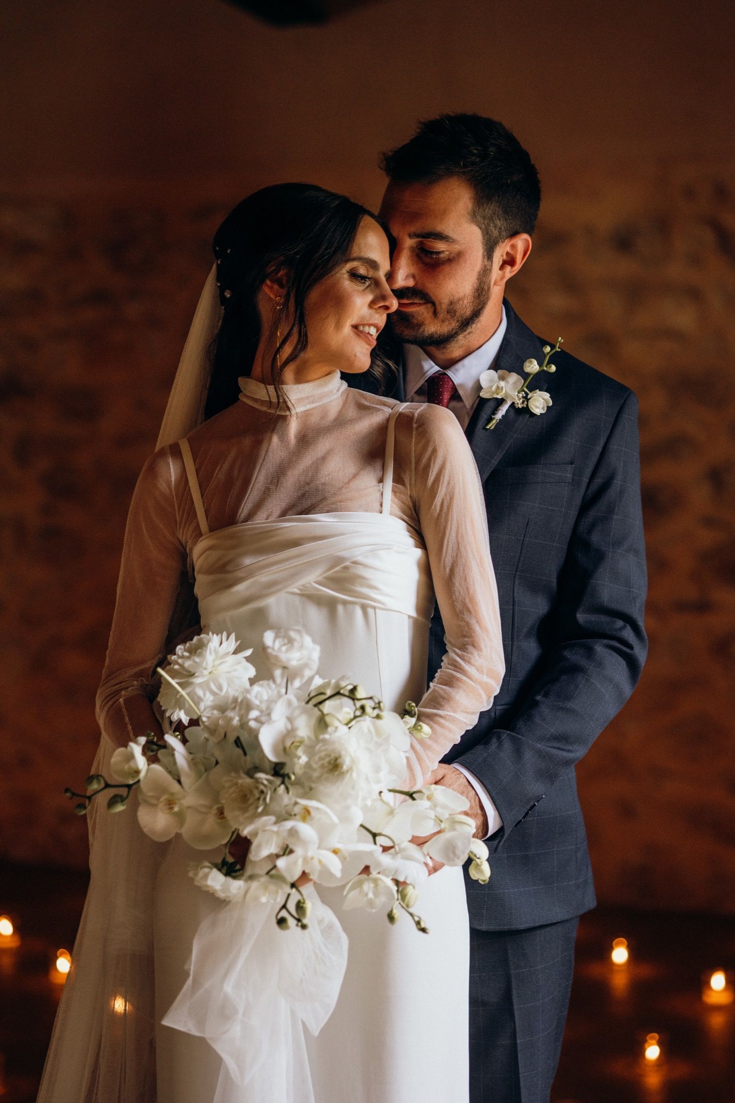 Romantic wedding couple embraces in candlelit venue with white floral bouquet and elegant bridal gown.