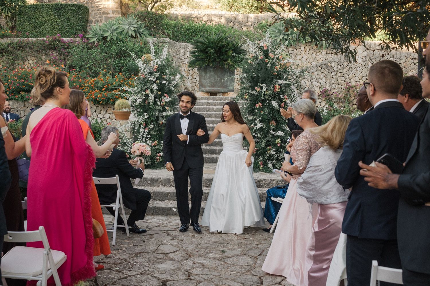 A wedding ceremony procession walks down stone steps in a garden setting with greenery and cliffs in the background.