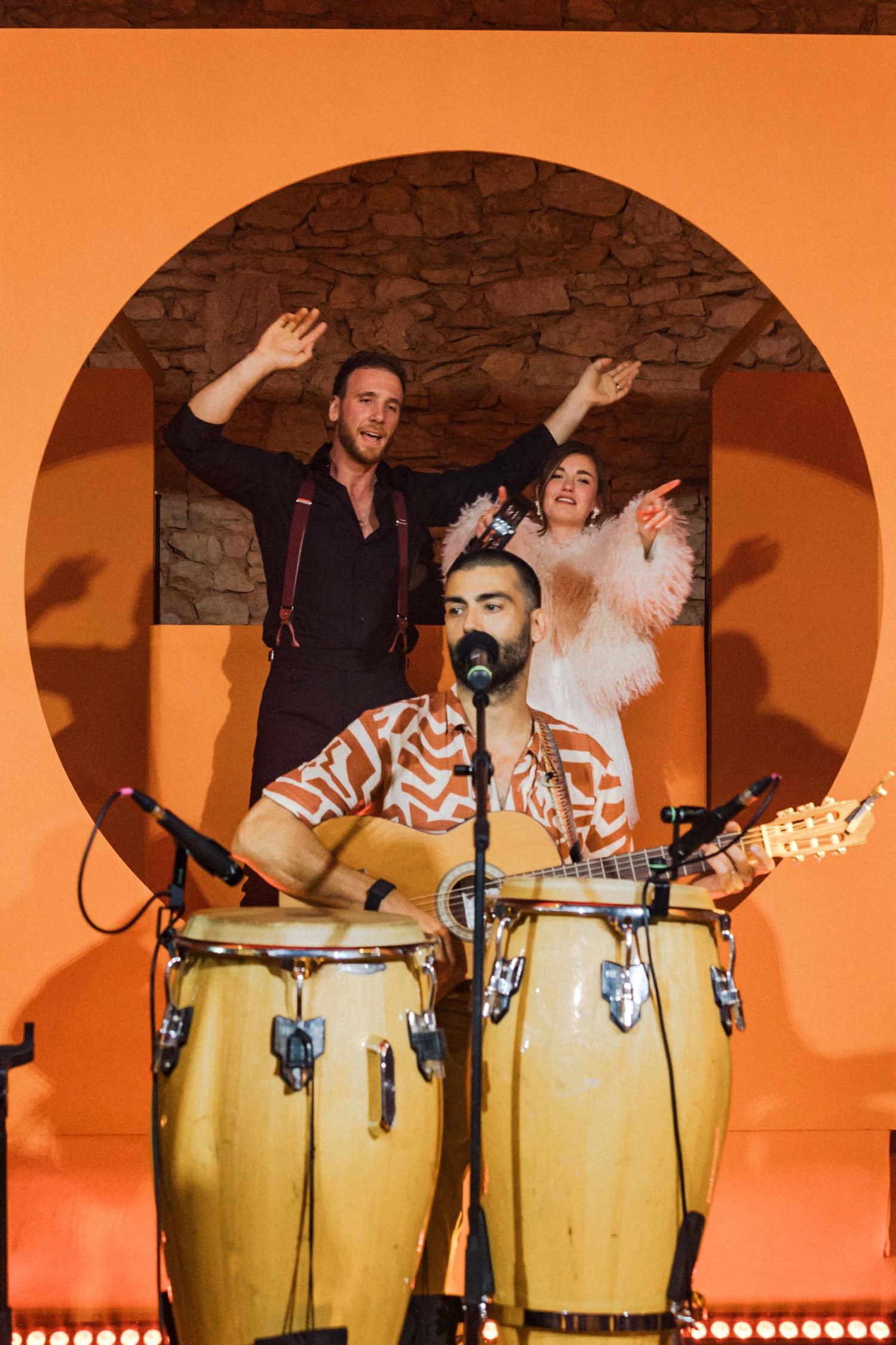 Musicians performing on stage with congas and percussion instruments against an orange wall with stone arch backdrop.