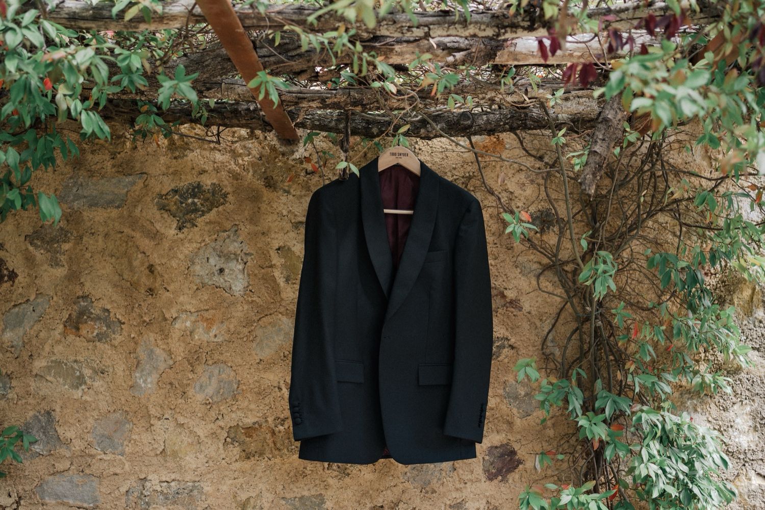 A black blazer hangs on a rustic stone wall surrounded by climbing green vines and foliage.