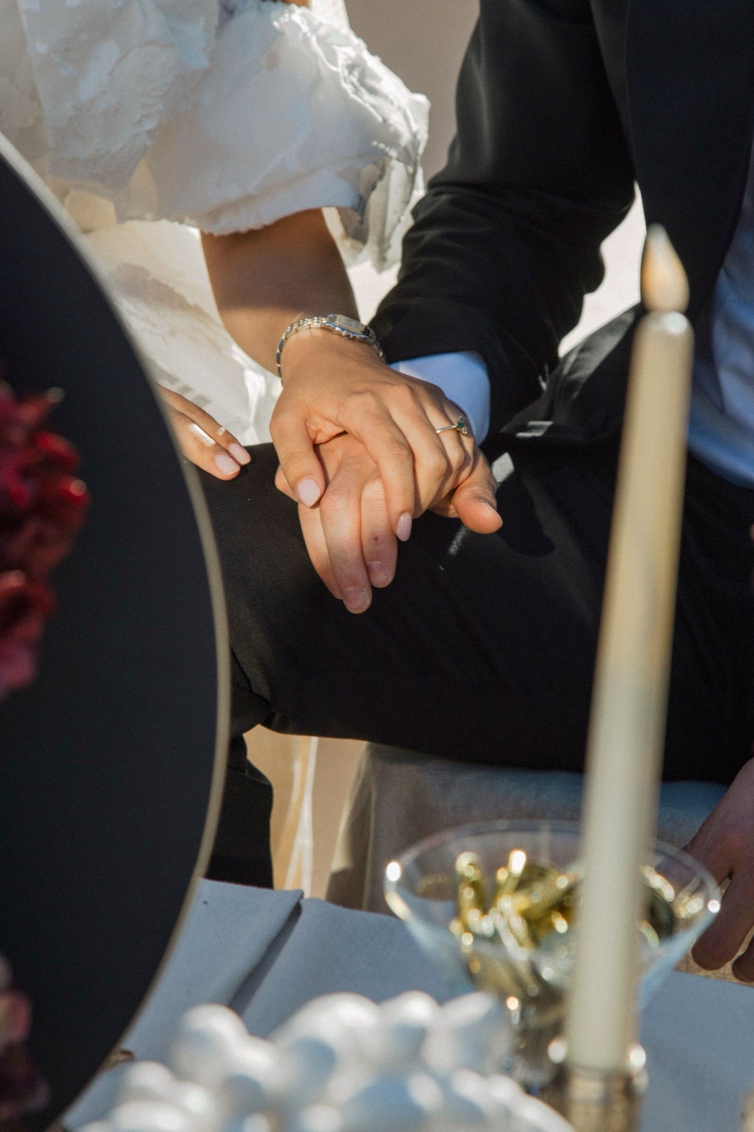 Close-up of bride and groom holding hands at their wedding reception table with candles and decor visible.