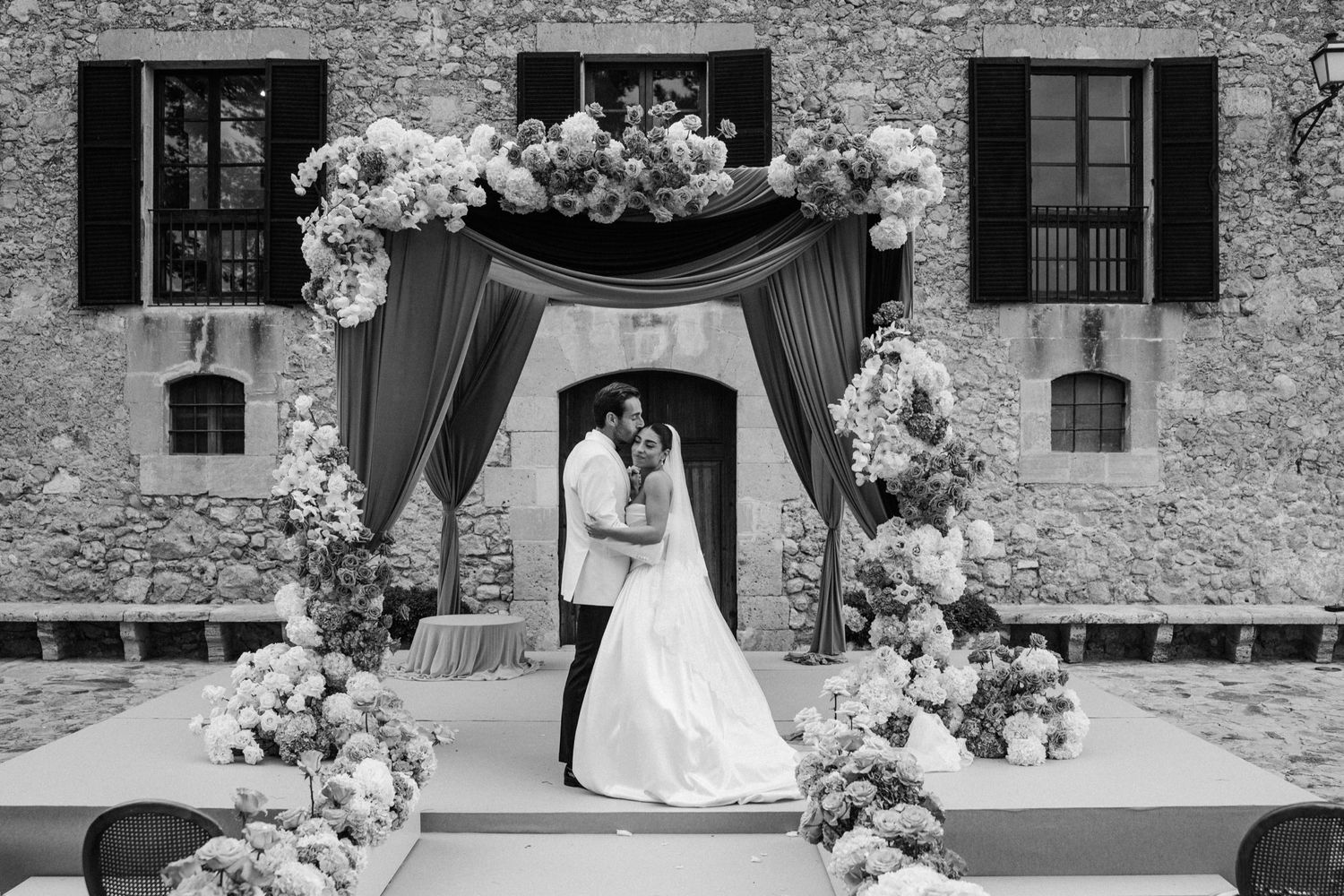 A romantic black and white wedding ceremony under a floral arch against a rustic stone wall with shuttered windows.