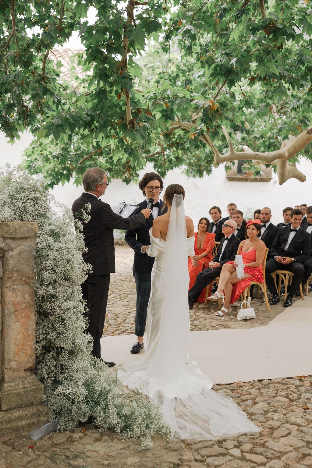 An outdoor wedding ceremony under a lush tree with guests seated on chairs watching the couple exchange vows.