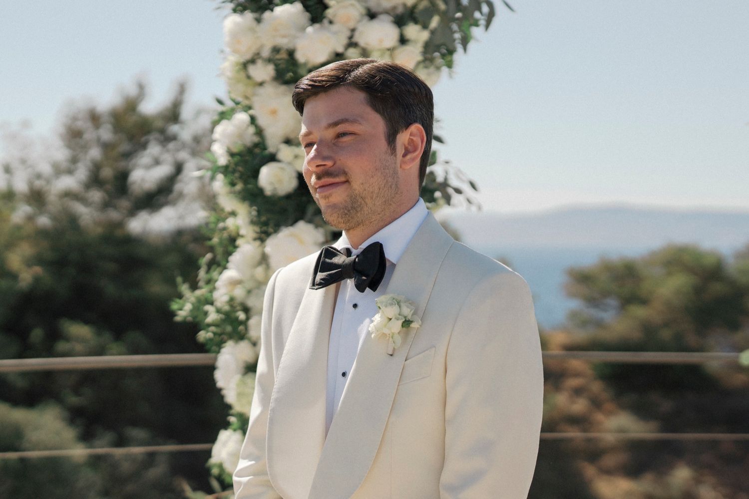 Groom in white tuxedo jacket and black bow tie standing at outdoor wedding altar with white floral arch.