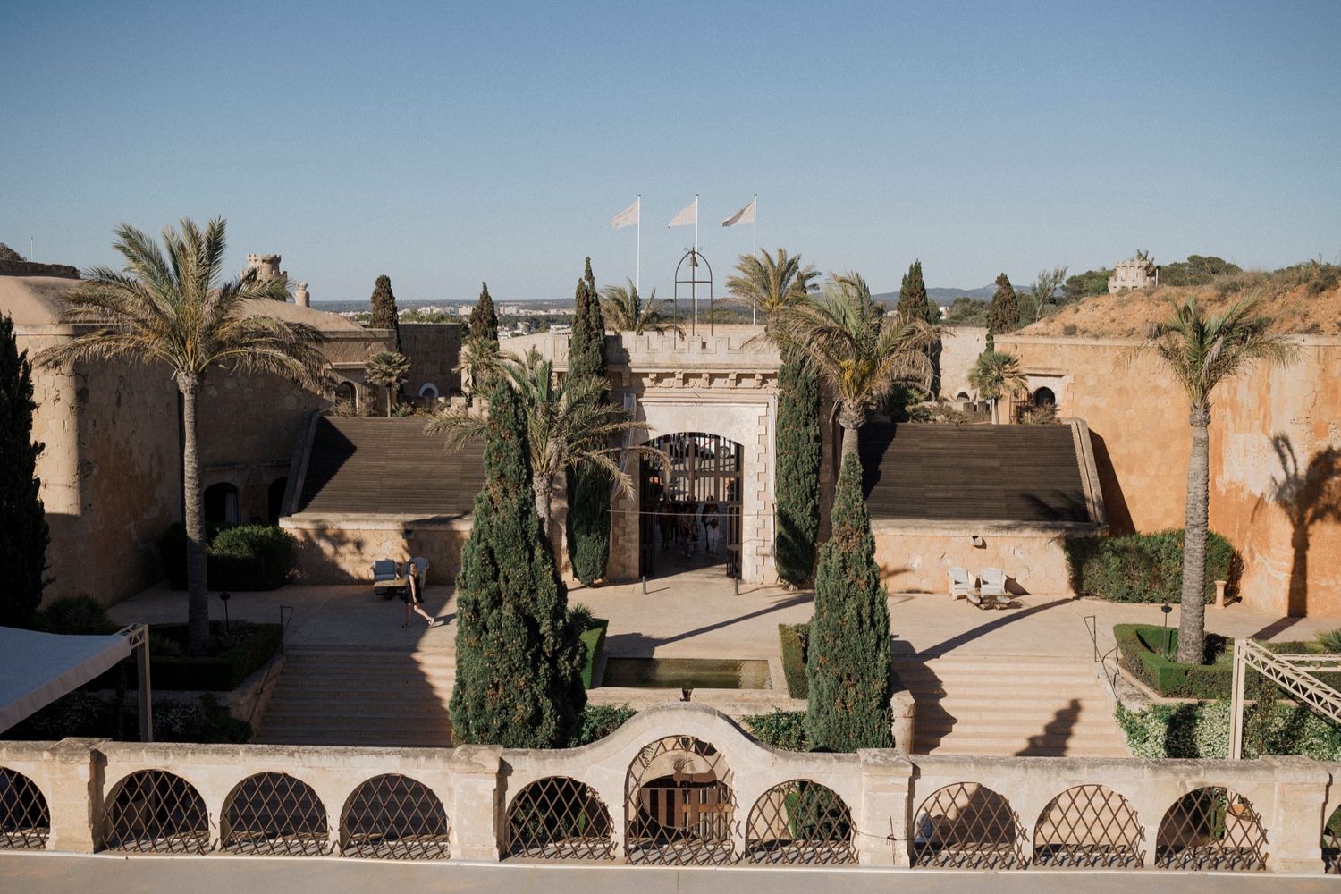 Aerial view of a historic fortress with palm trees, arched walkways, and cypress trees under a clear blue sky.