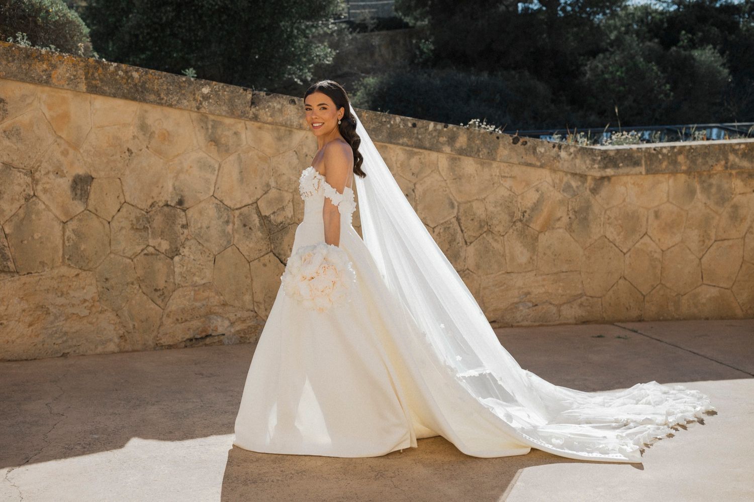 Bride in elegant white ball gown with long veil poses gracefully outdoors against a stone wall.