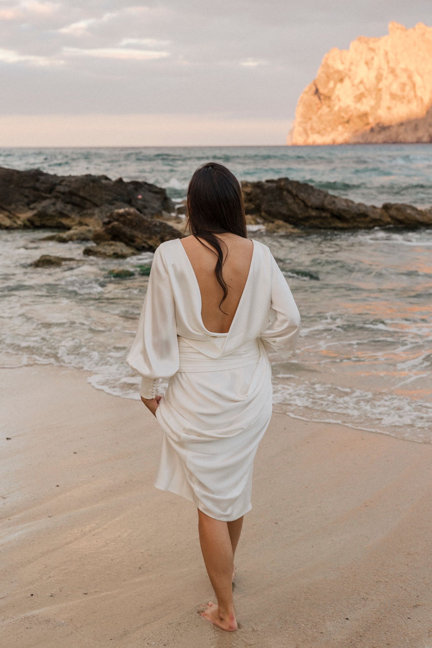 Woman in flowing white dress walks barefoot on beach at sunset with dramatic cliffside backdrop.