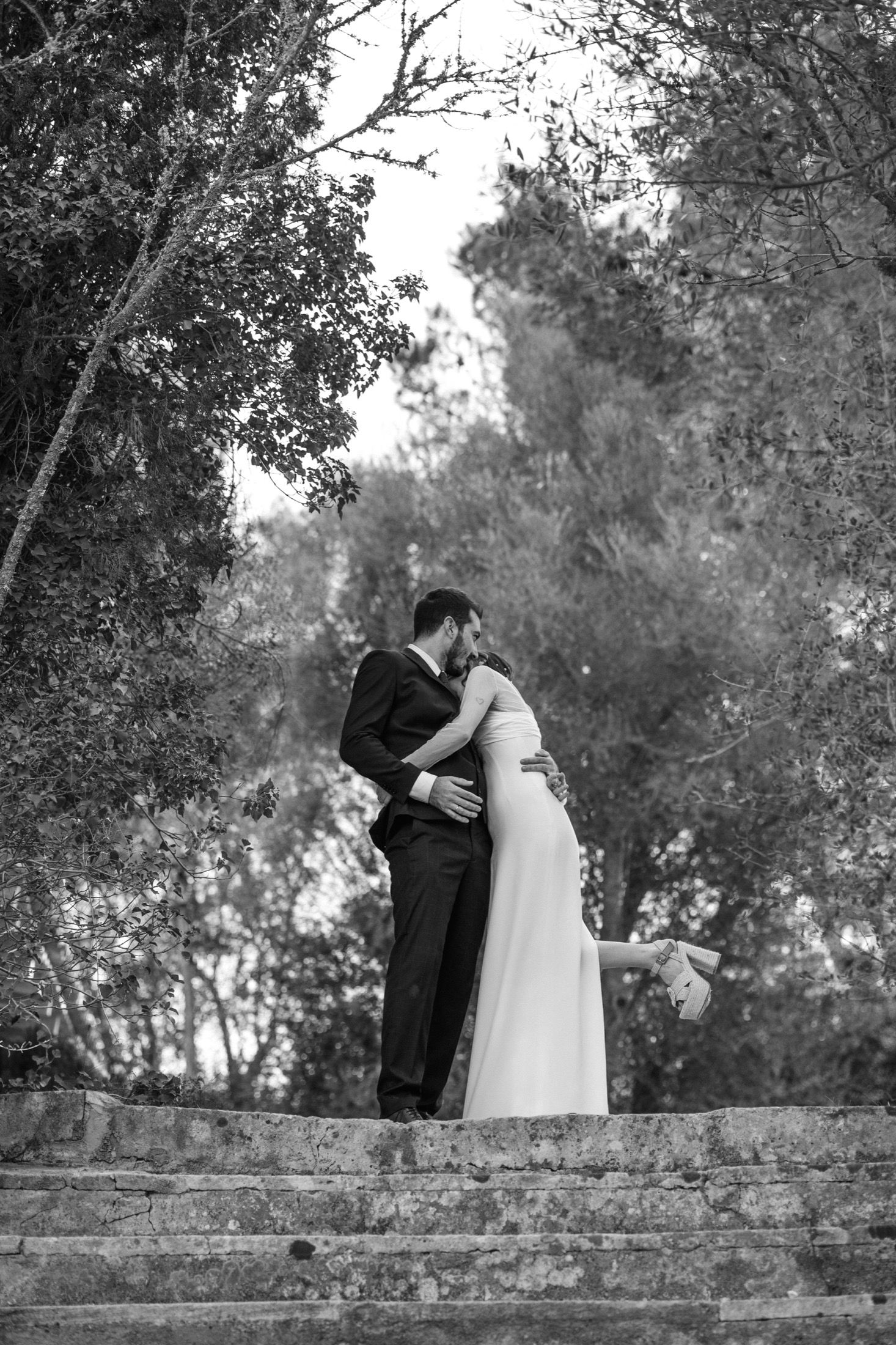 Black and white wedding photos show romantic moments between a couple embracing on a rustic stone wall in a garden setting.