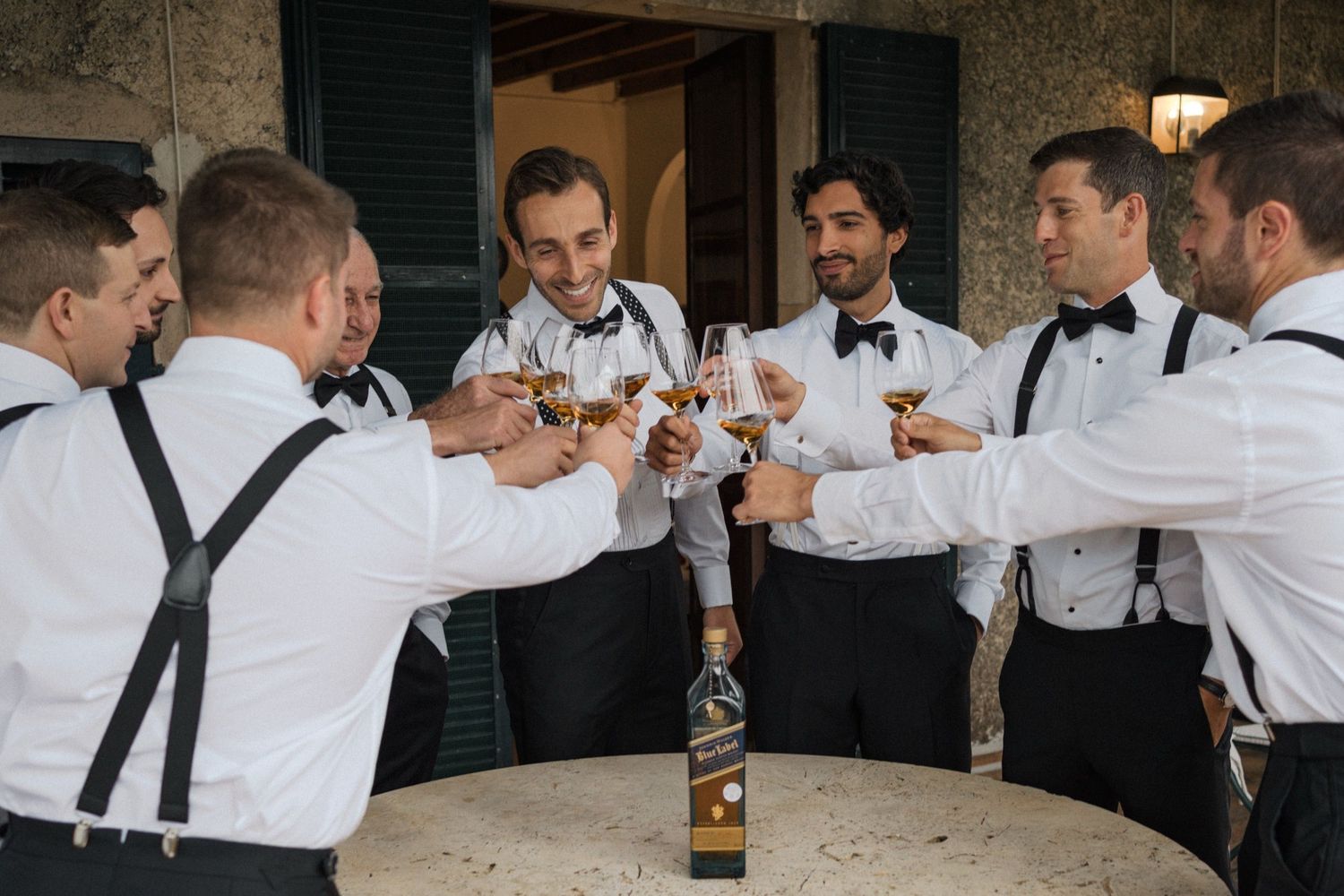 A group of groomsmen in black and white attire raise their drinks in celebration at a wedding reception.