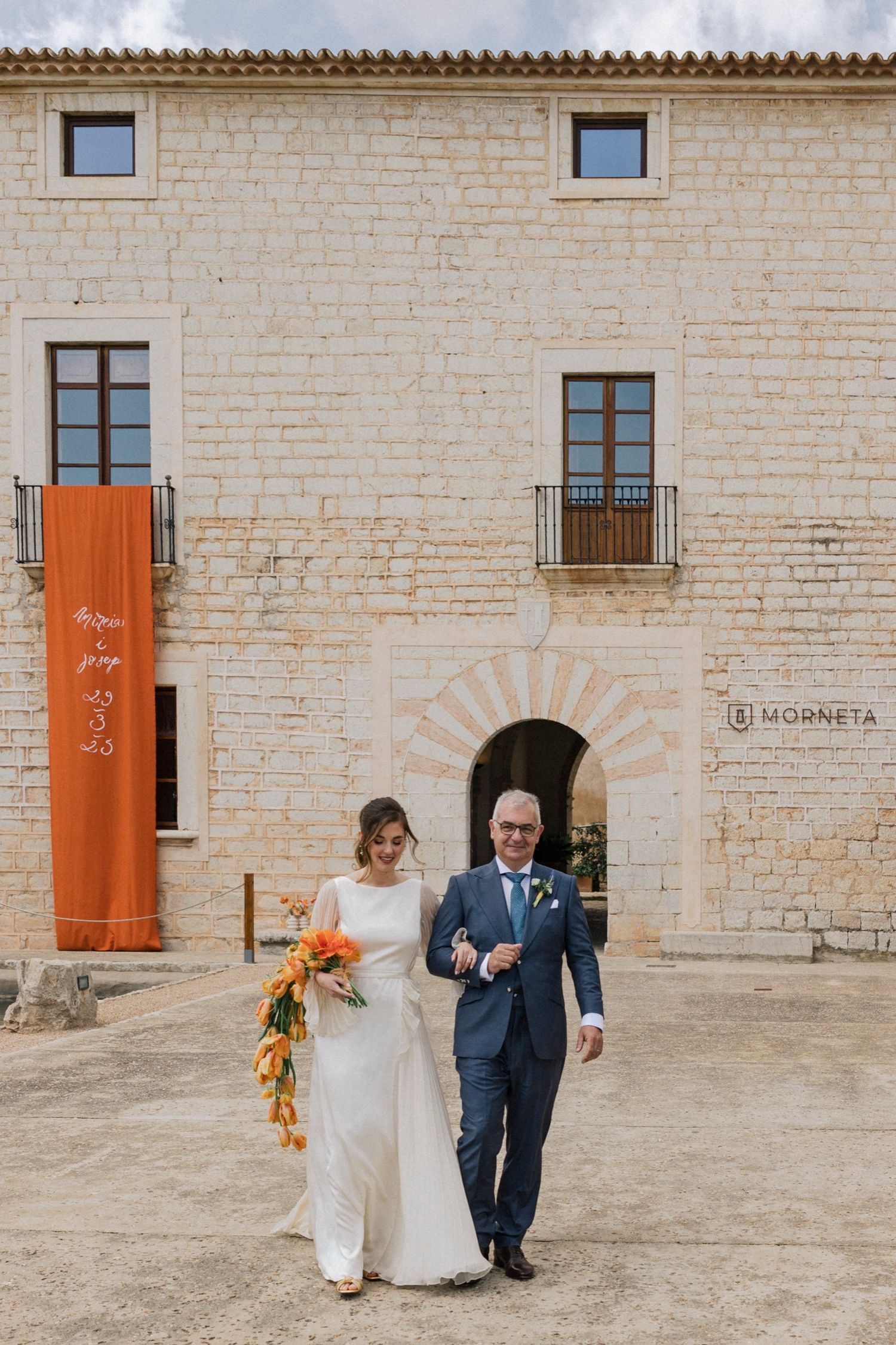 A wedding couple walks together outside a rustic stone building with orange banners and arched windows.