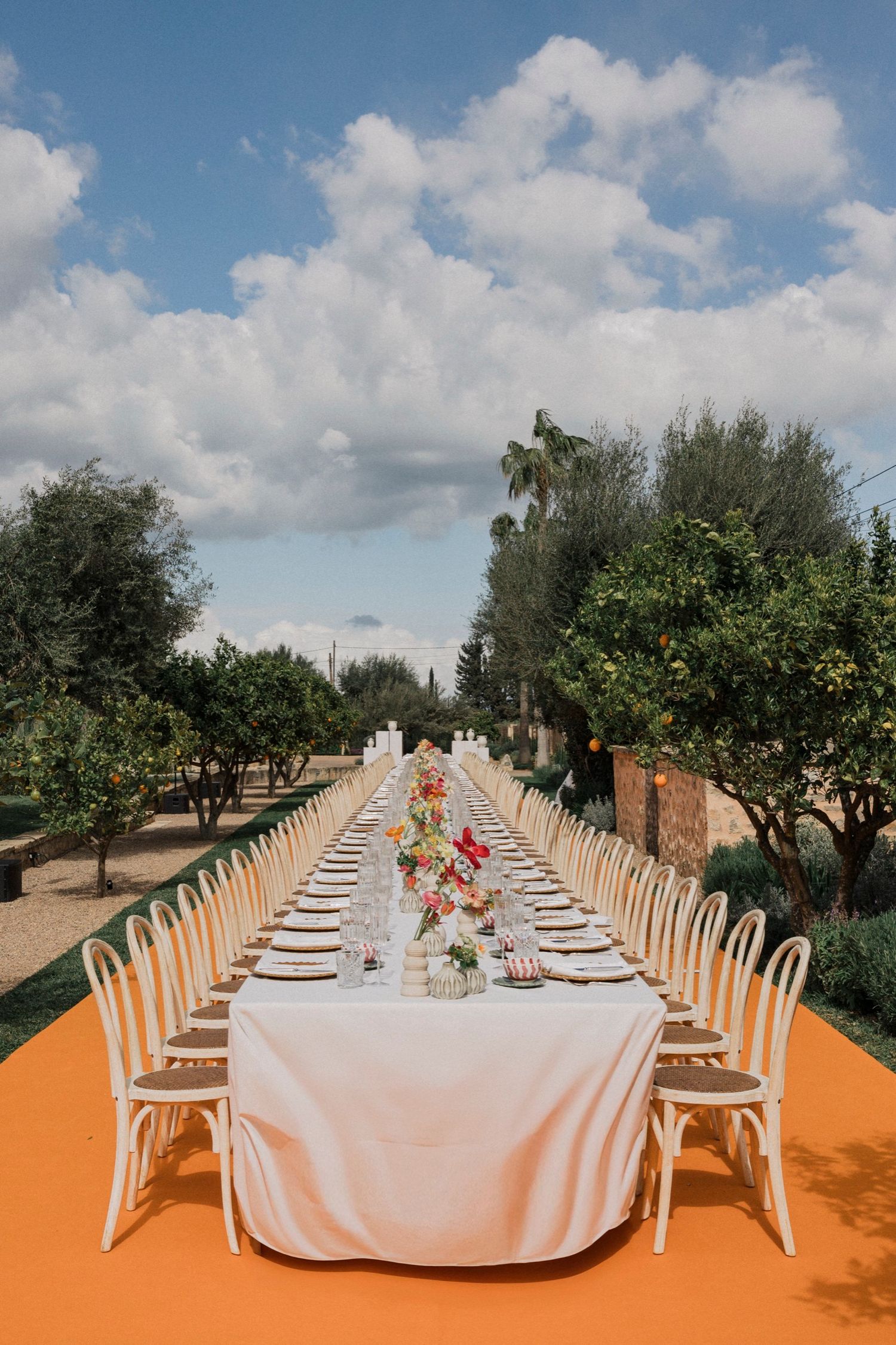 Long dining table with white linens and chairs set up outdoors in an orange-floored venue with olive trees.