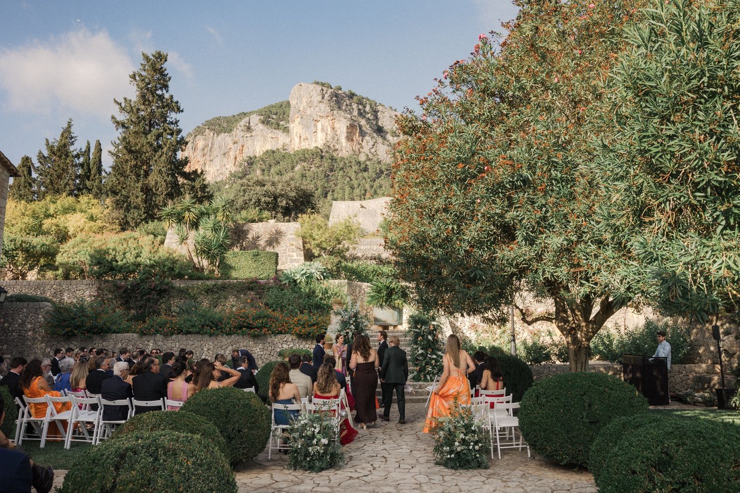 Outdoor wedding ceremony setup with white chairs in a garden against a backdrop of cliffs and greenery.