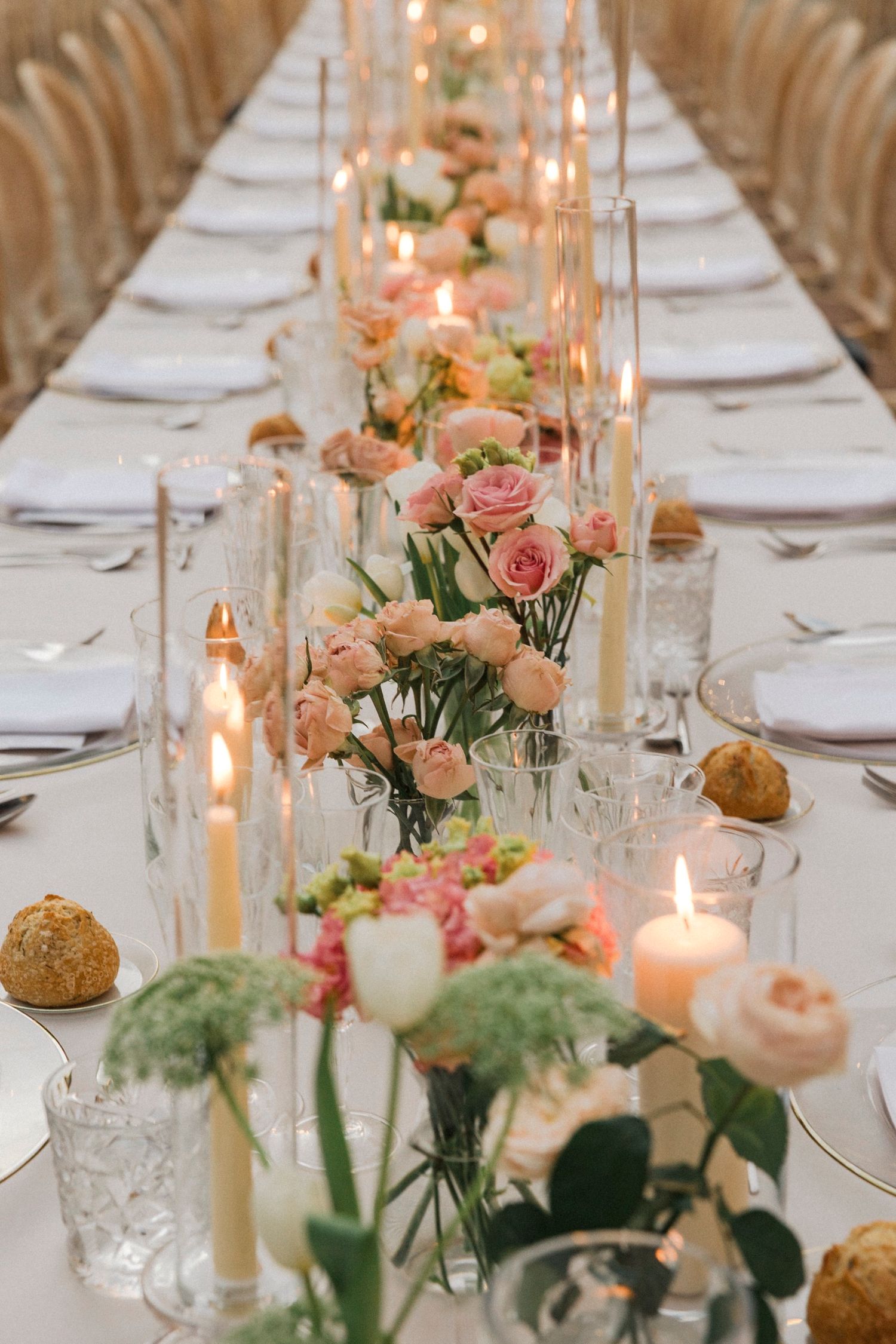 Elegant wedding table setting with pink roses, taper candles, crystal glassware and white linens in a rustic venue.