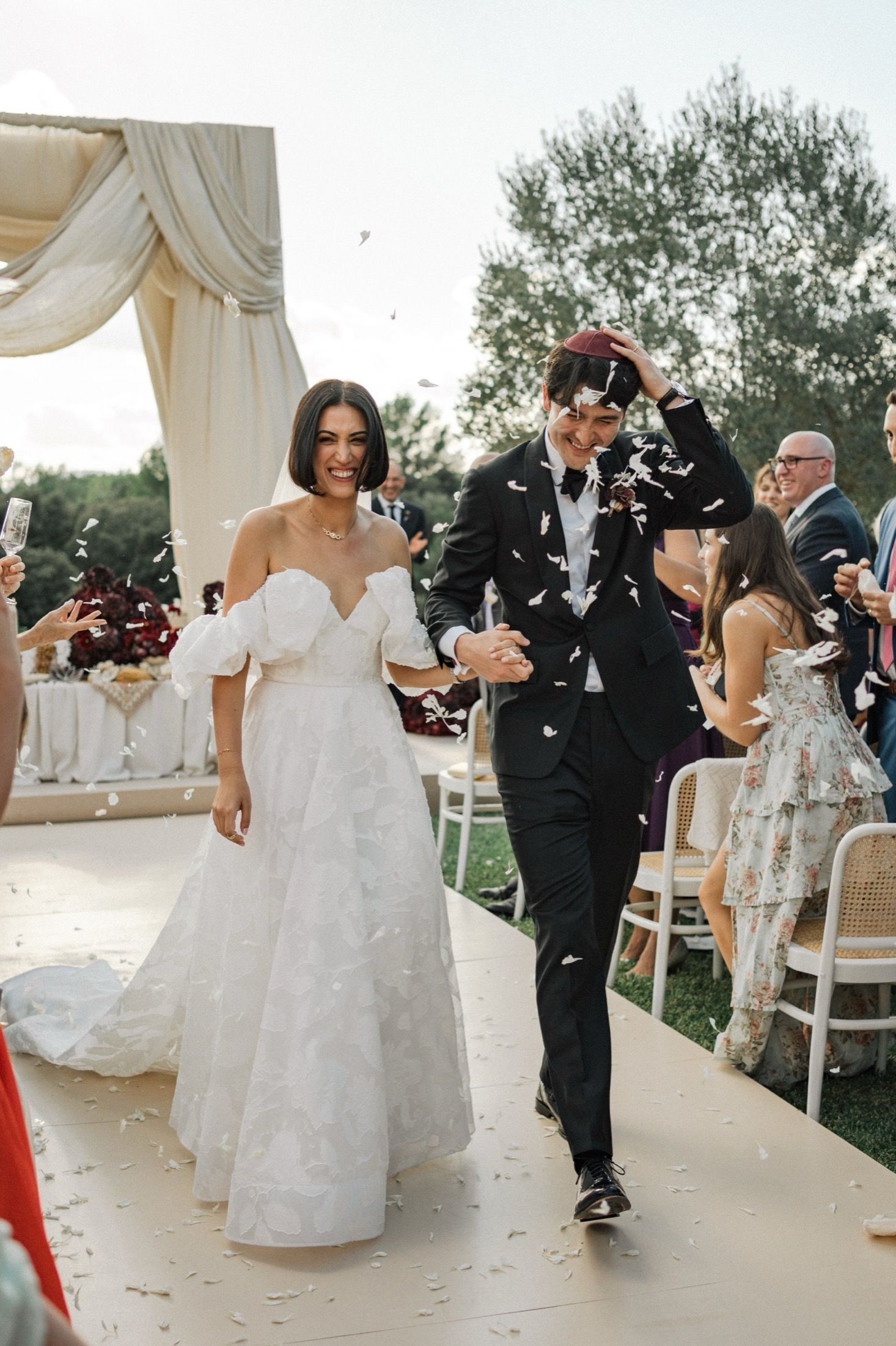 Joyful newlyweds walk down outdoor aisle under white draped arch while guests shower them with confetti.