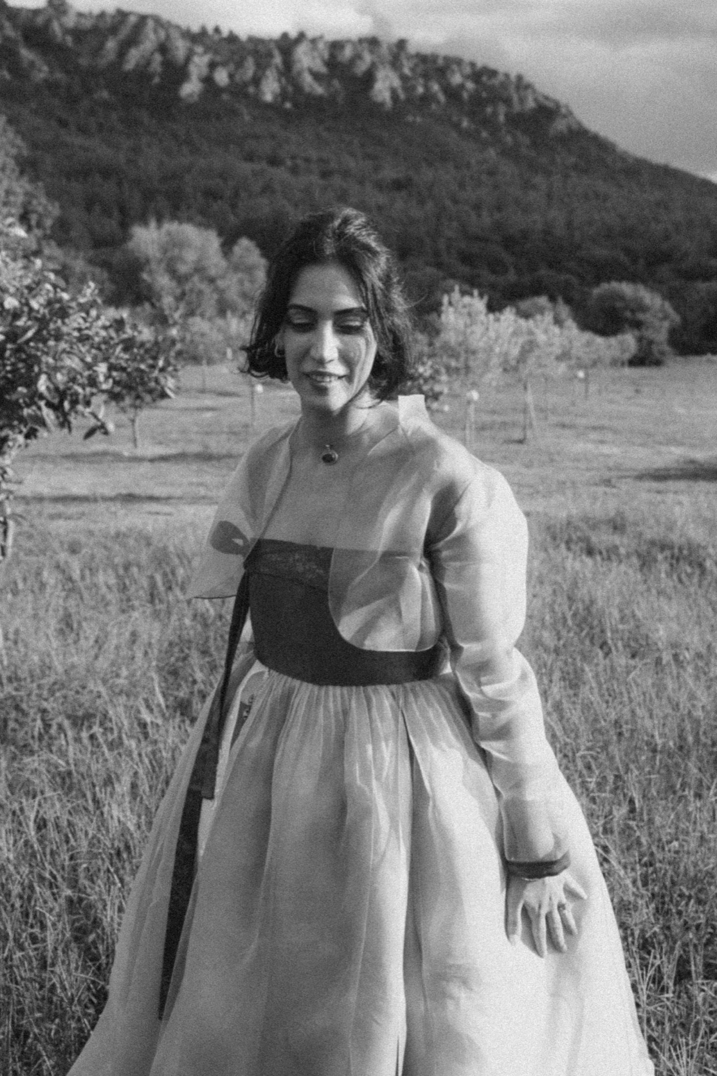 Black and white photo of someone in a traditional dress standing in a field with mountains in the background.