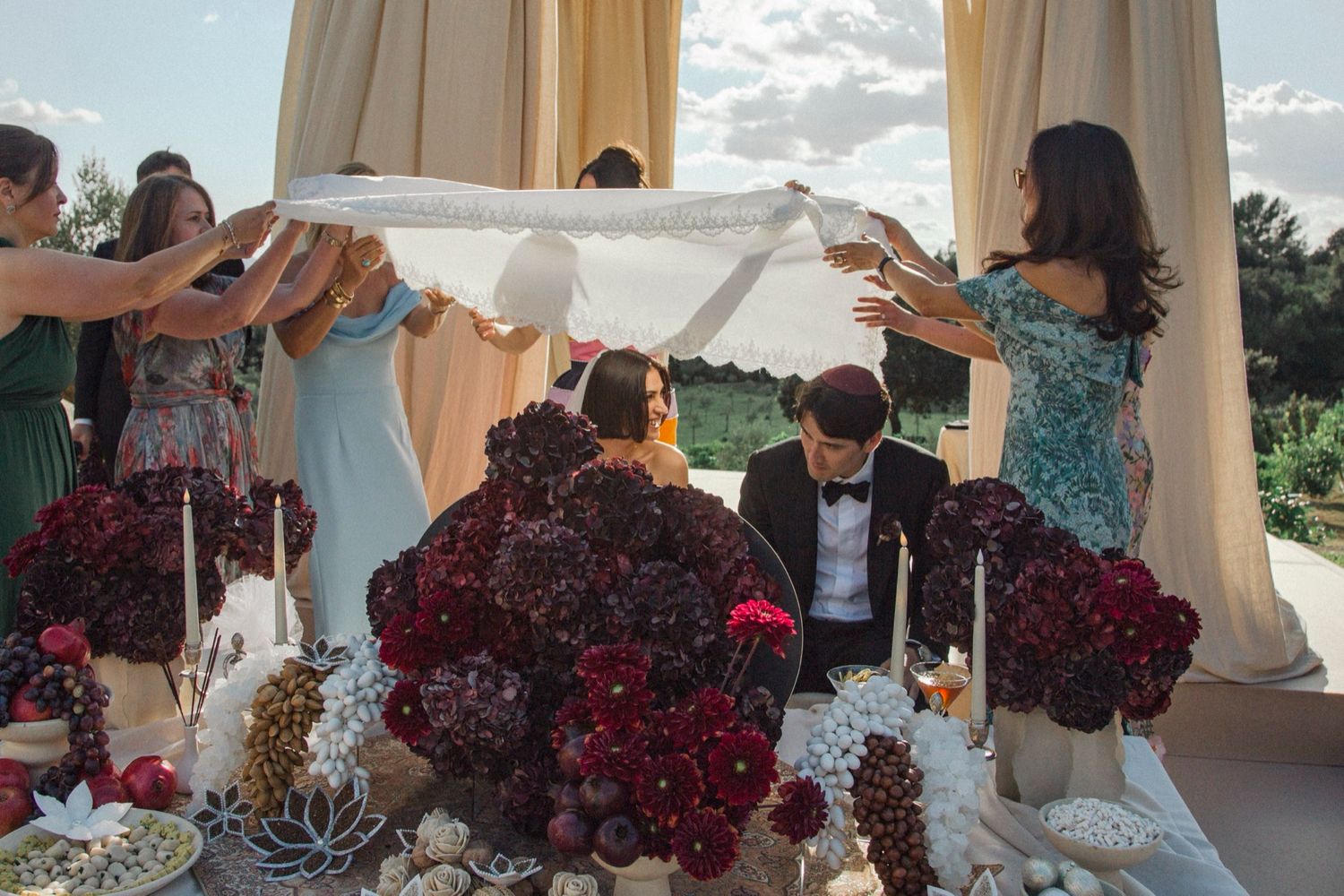 Wedding party celebrates with burgundy bouquets under a white tent during an outdoor reception.