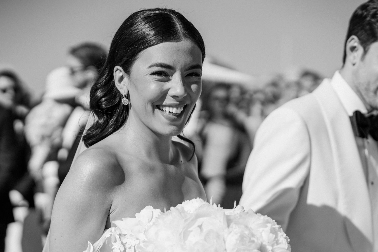 Radiant bride smiling and holding a floral bouquet in a black and white outdoor wedding photo.