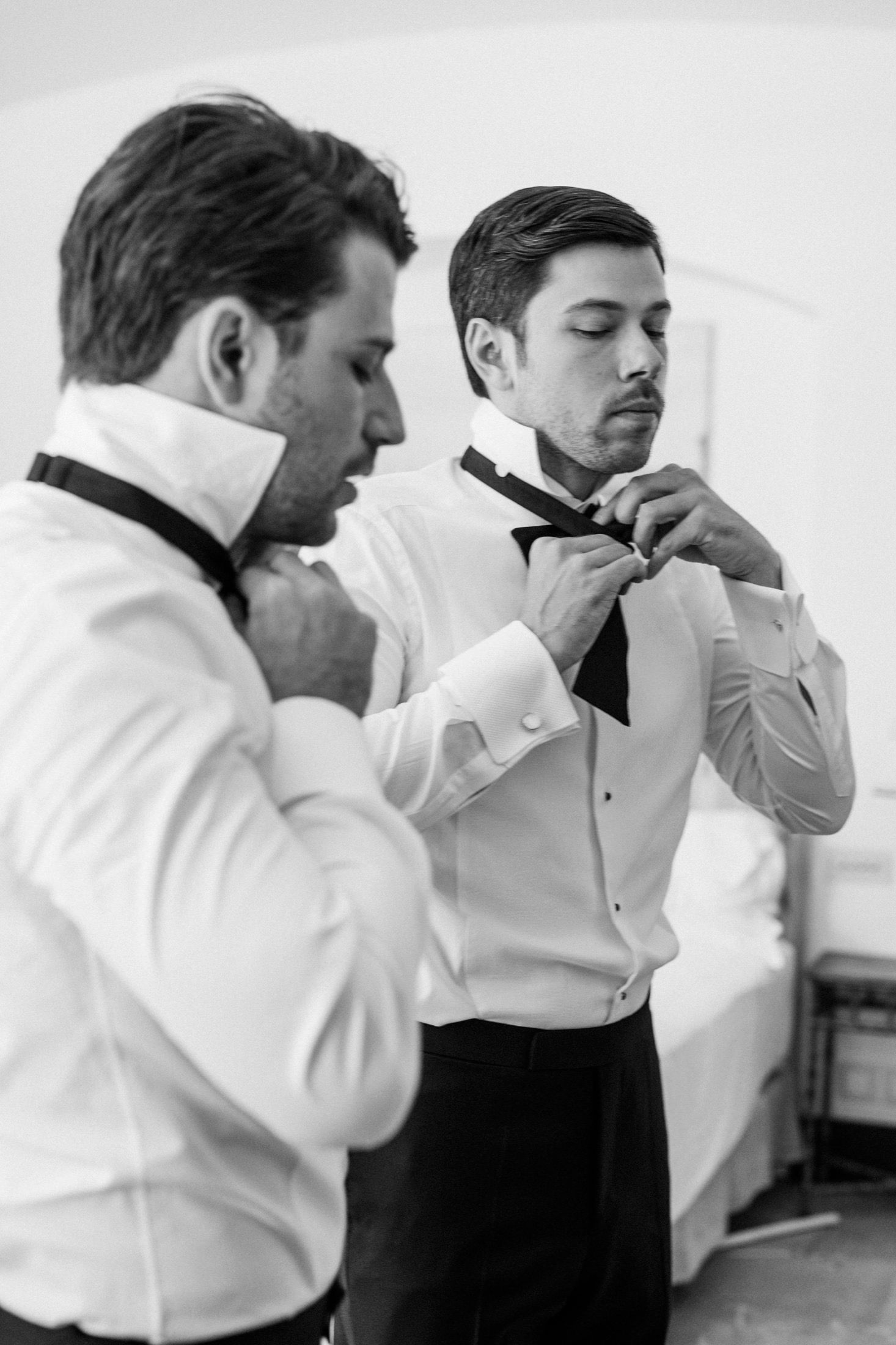 Two groomsmen in white tuxedo shirts adjusting black bow ties while preparing for a wedding ceremony.