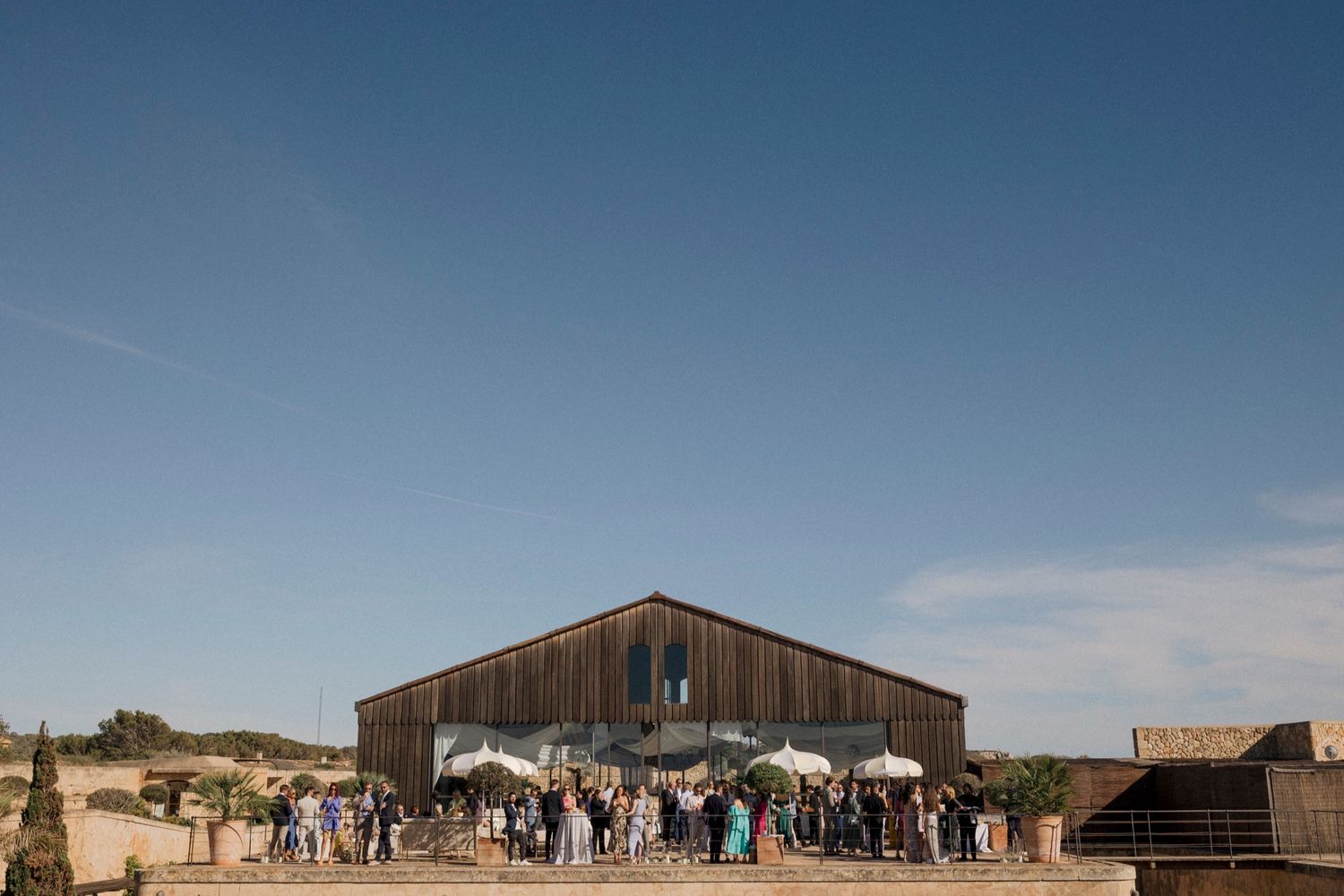 Guests gather outside a rustic wooden barn wedding venue under a clear blue sky.