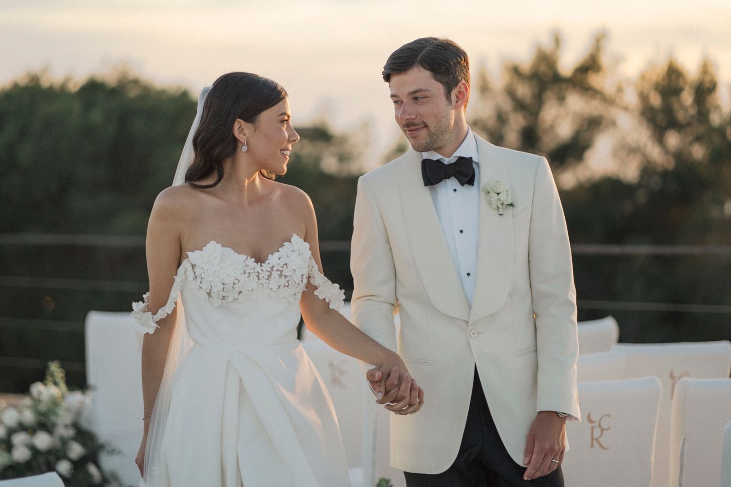 Bride in strapless white gown and groom in ivory tuxedo hold hands at outdoor wedding ceremony at sunset.