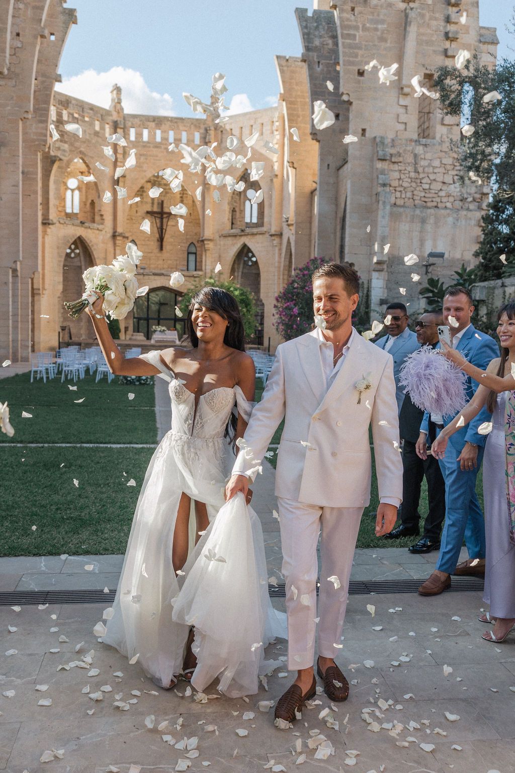 Wedding party celebrates with flower petals at historic stone church courtyard.