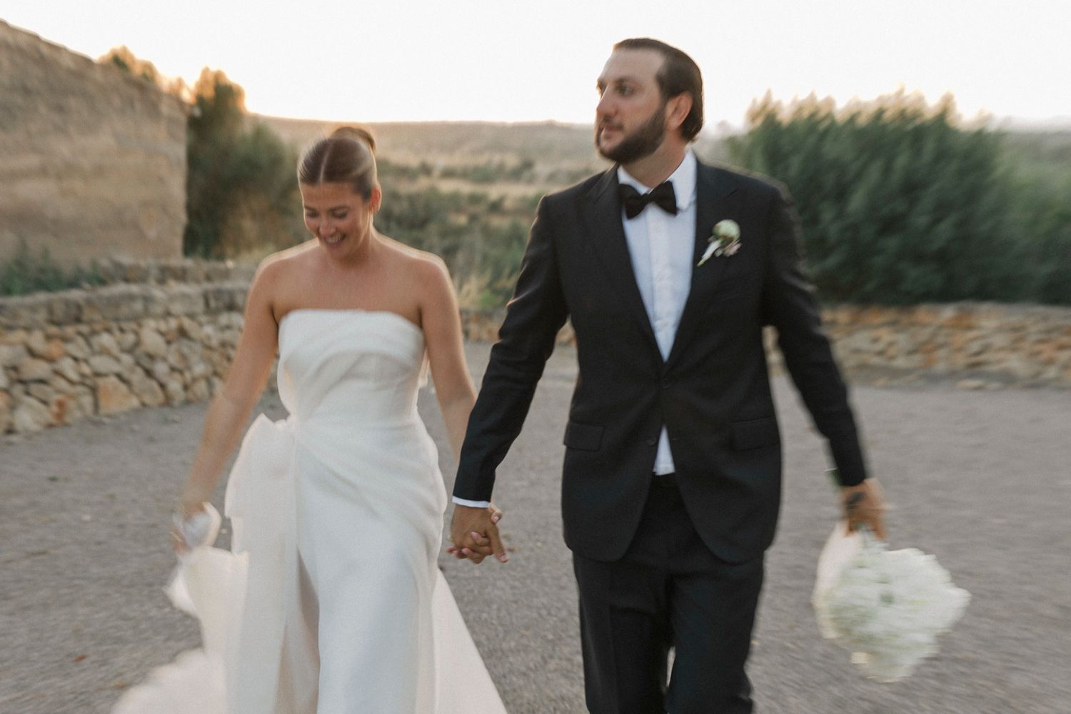 Elegant couple walking through a rustic stone courtyard in formal wedding attire.