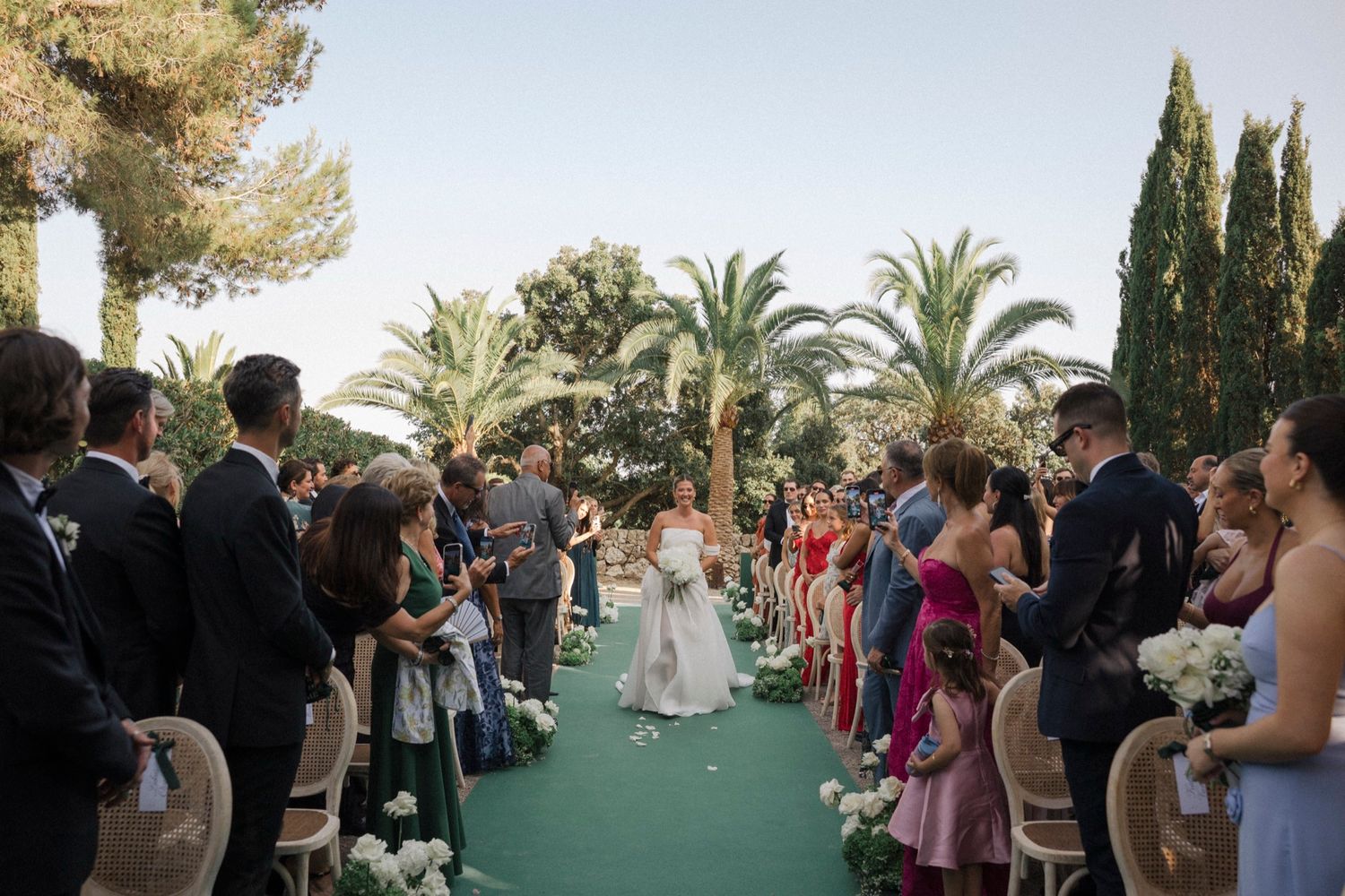 Wedding ceremony guests line a green carpet aisle surrounded by palm trees and cypress in an outdoor garden setting.