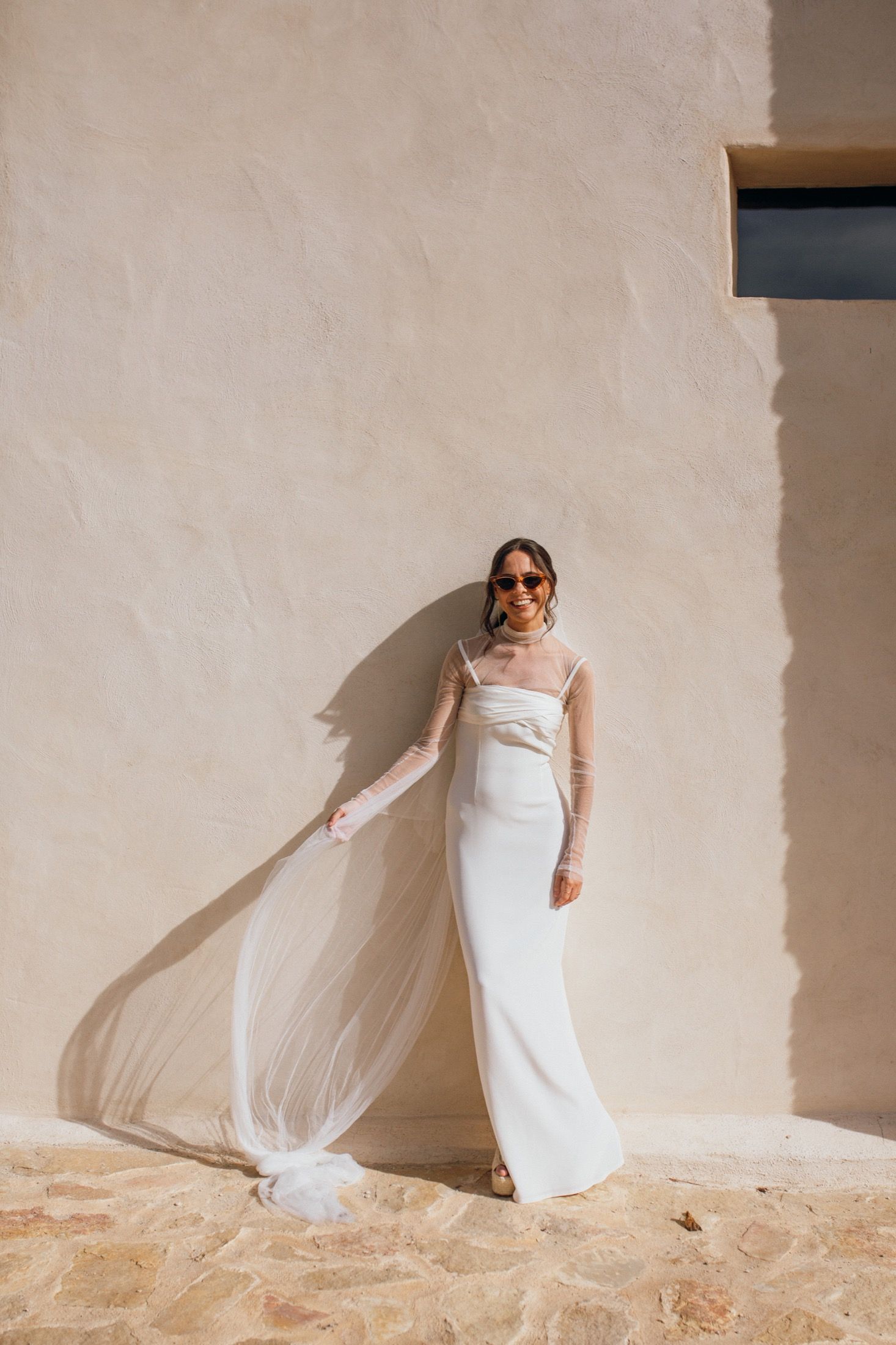 A bride in an elegant white gown with flowing veil poses against a textured beige wall in natural sunlight.