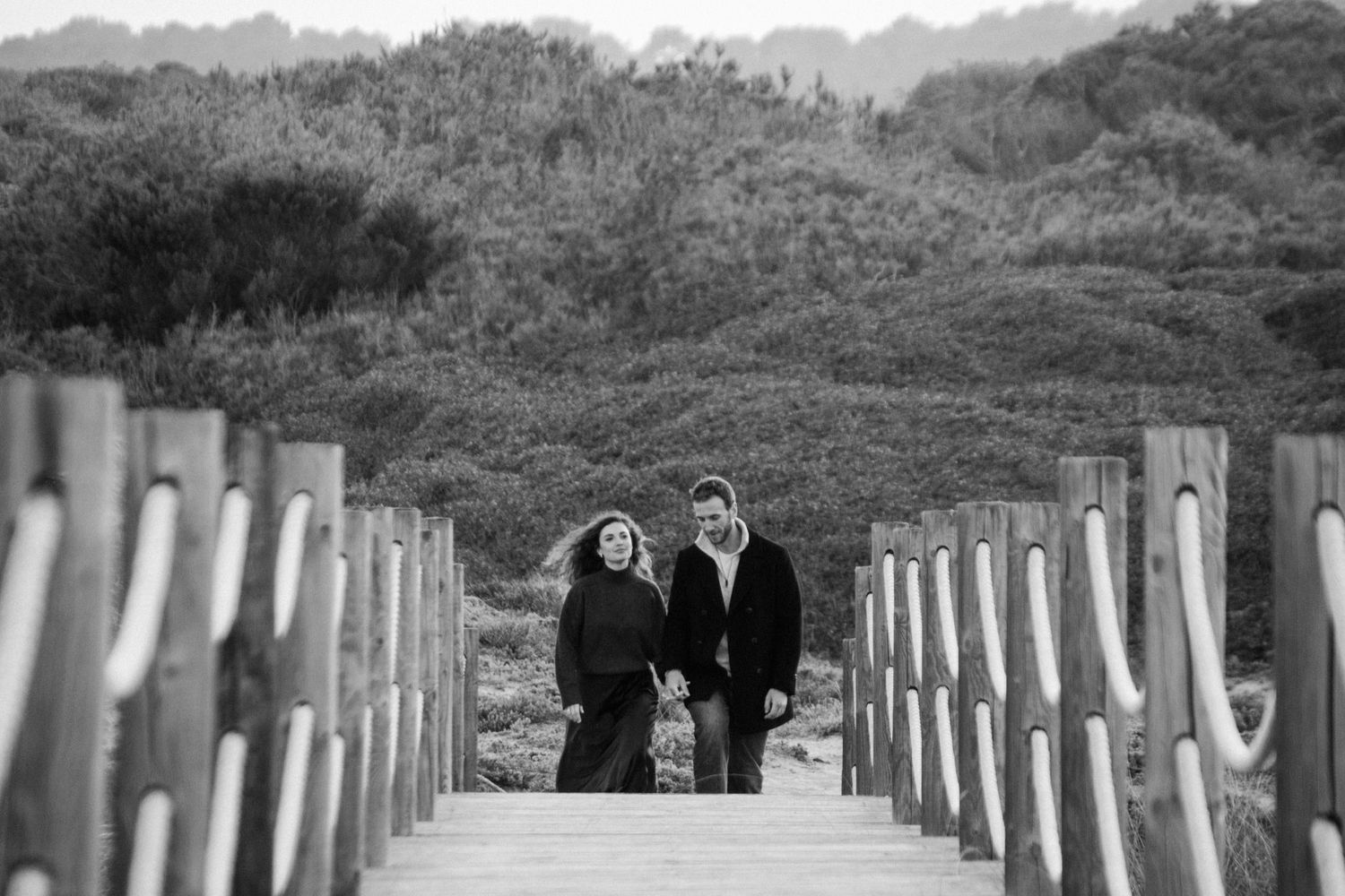A couple walks together on a wooden boardwalk path lined with white posts in a black and white photograph.